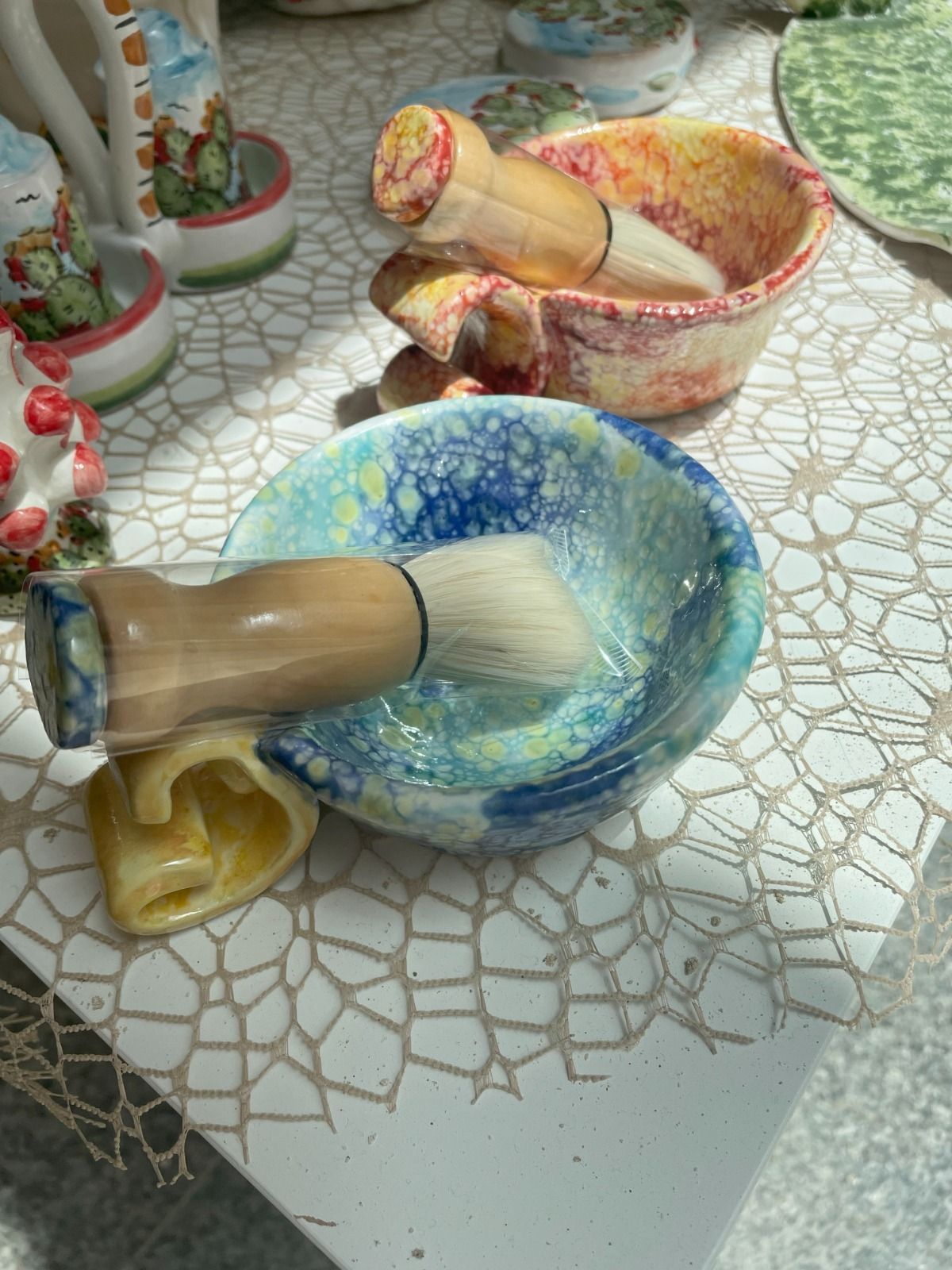 Ciotola da barba con pennello. La ciotola è blu, gialla e bianca, e sullo sfondo ci sono altri oggetti in ceramica.