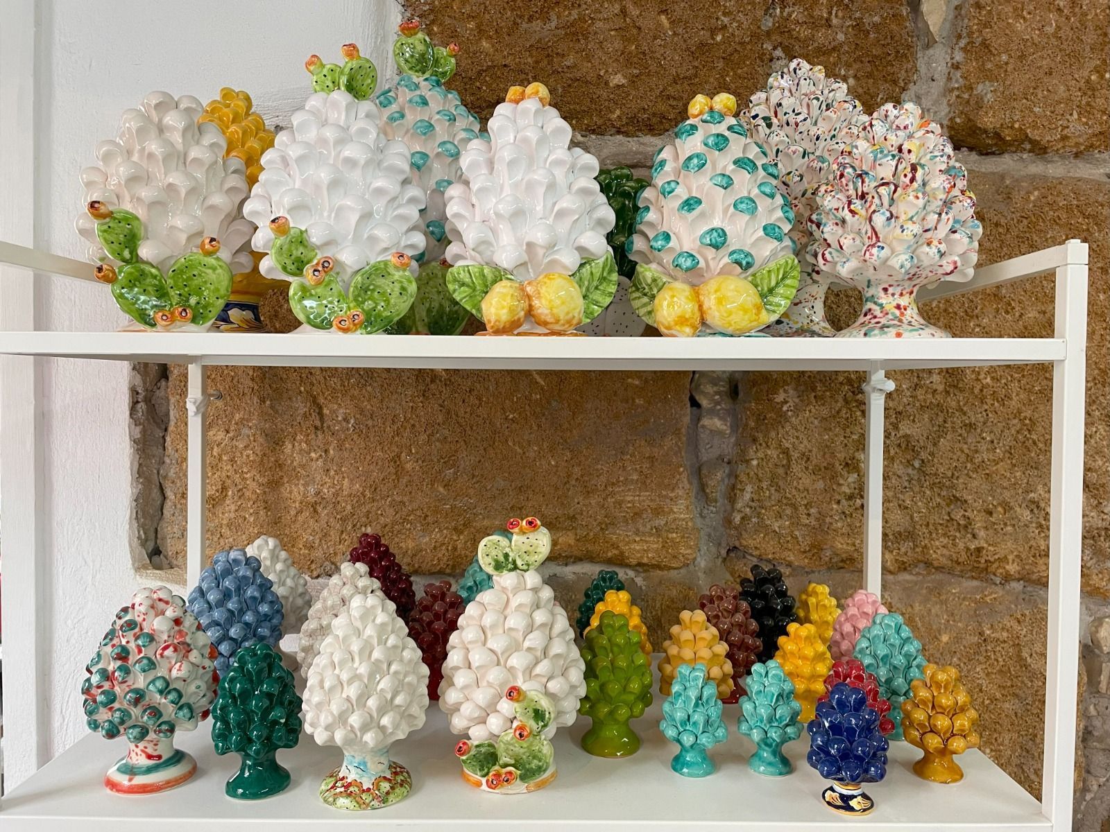 Scaffali con sculture di pigne in ceramica colorate, appoggiate a un muro di pietra.