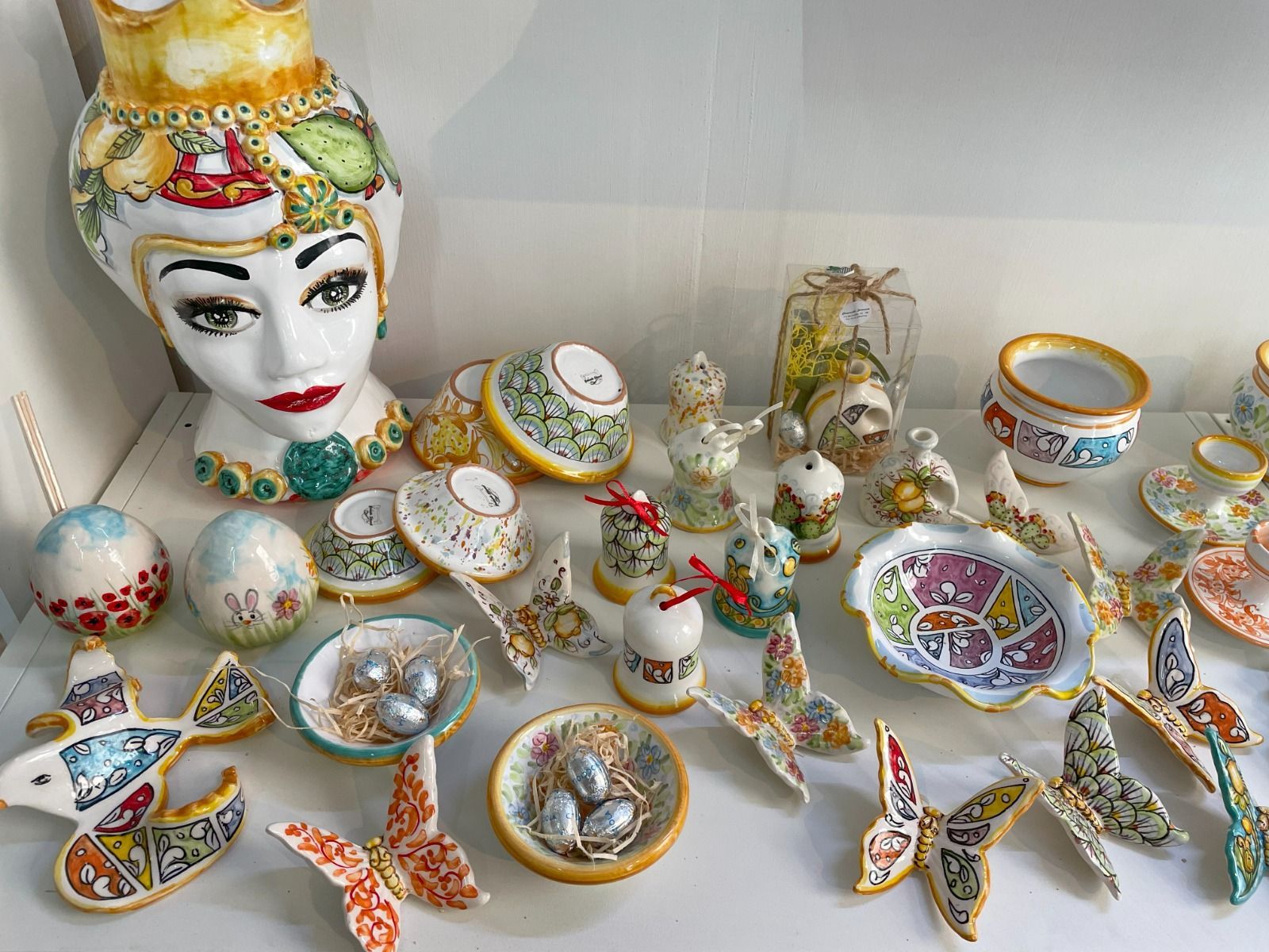 Esposizione di opere d'arte in ceramica: volti colorati, farfalle, ciotole, uova e vasi, con motivi e dettagli intricati, su uno scaffale.