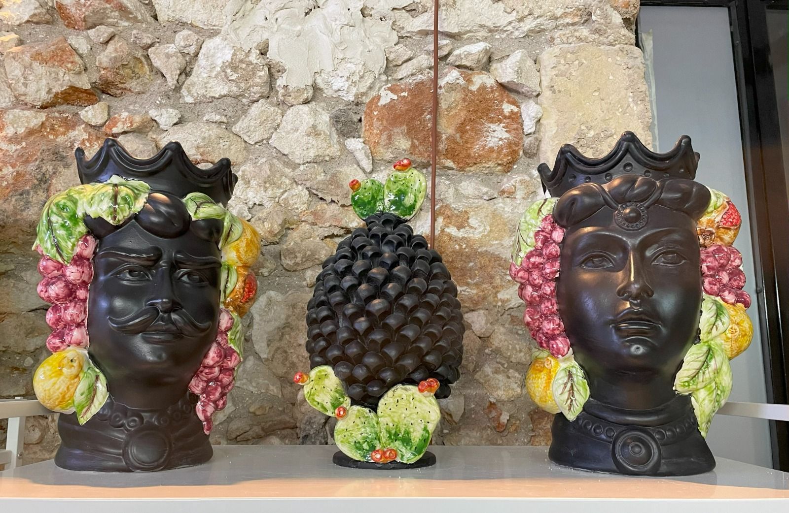 Tre teste di ceramica scura con decorazioni di frutta sono poste davanti a un muro di pietra; al centro c'è un oggetto a forma di ananas.