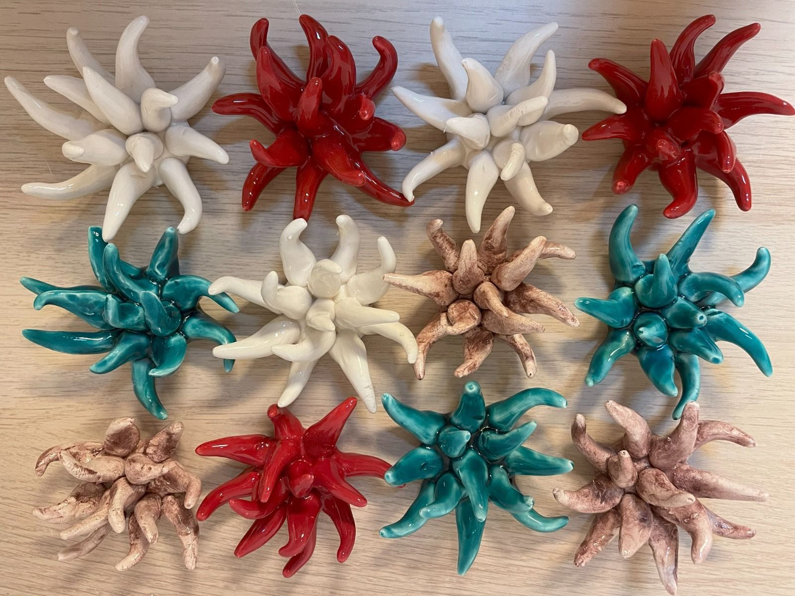 Sculture in ceramica a forma di stella nei colori bianco, rosso, verde acqua e marrone chiaro, disposte su una superficie di legno chiaro.