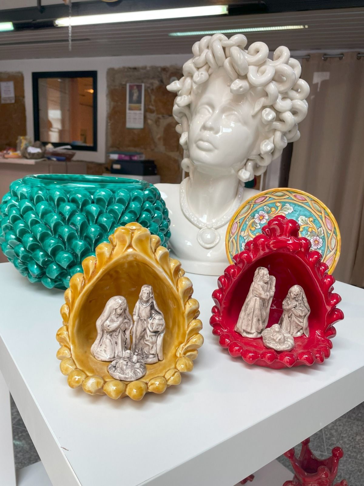 Esposizione di statuette in ceramica: busti, vasi e presepi dai colori vivaci su una superficie bianca.