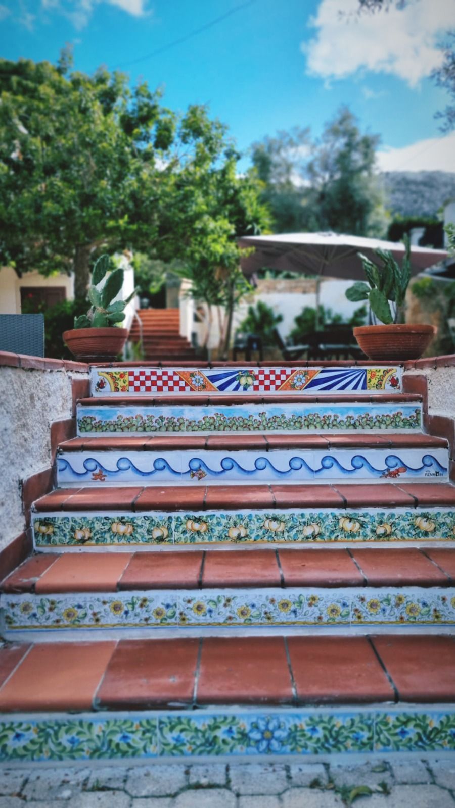 Scala con piastrelle decorative in un giardino, gradini in terracotta, cielo azzurro, alberi verdi sullo sfondo.