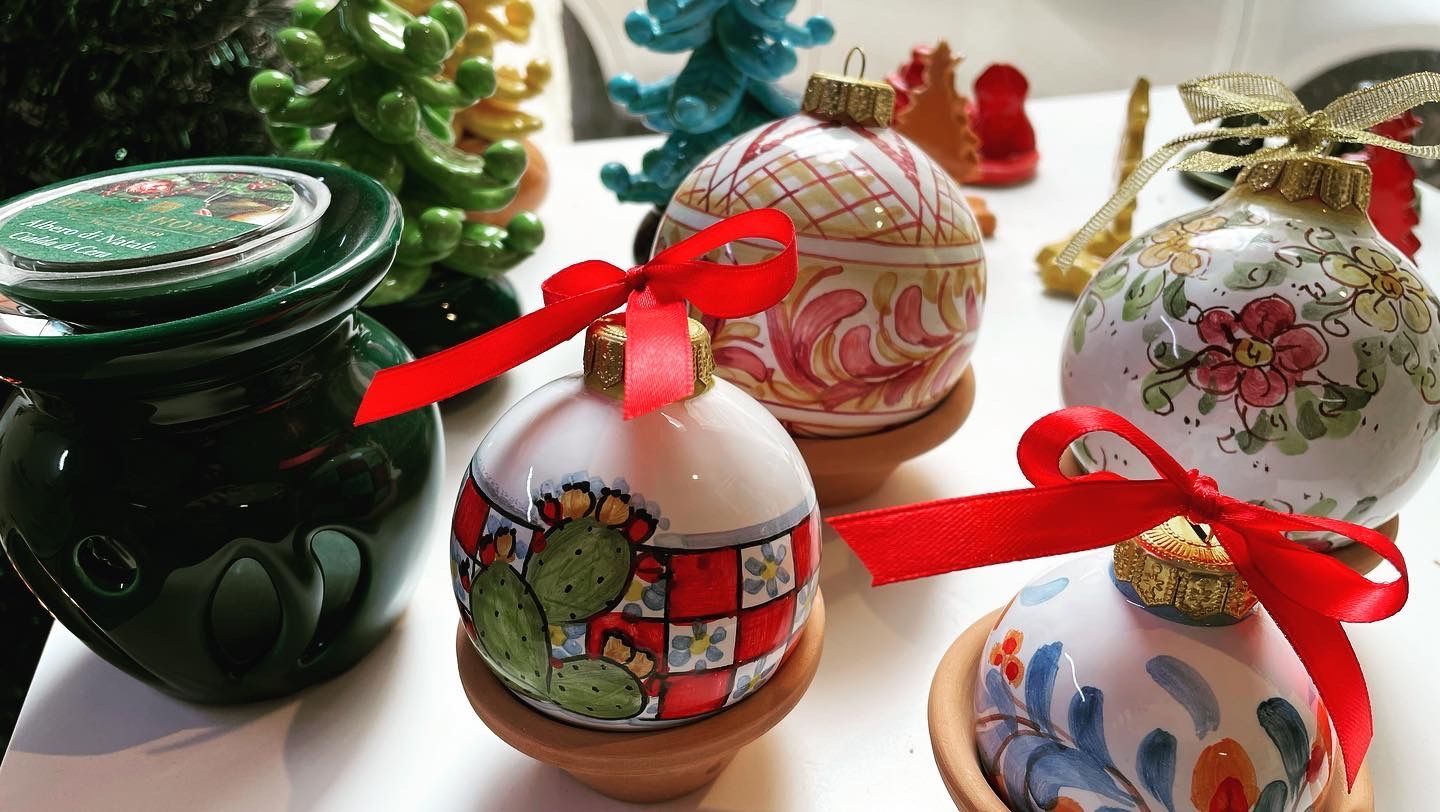 Decorazioni natalizie, alcune con fiocchi rossi e motivi colorati dipinti, su supporti. Vasi verdi e alberi di ceramica sullo sfondo.