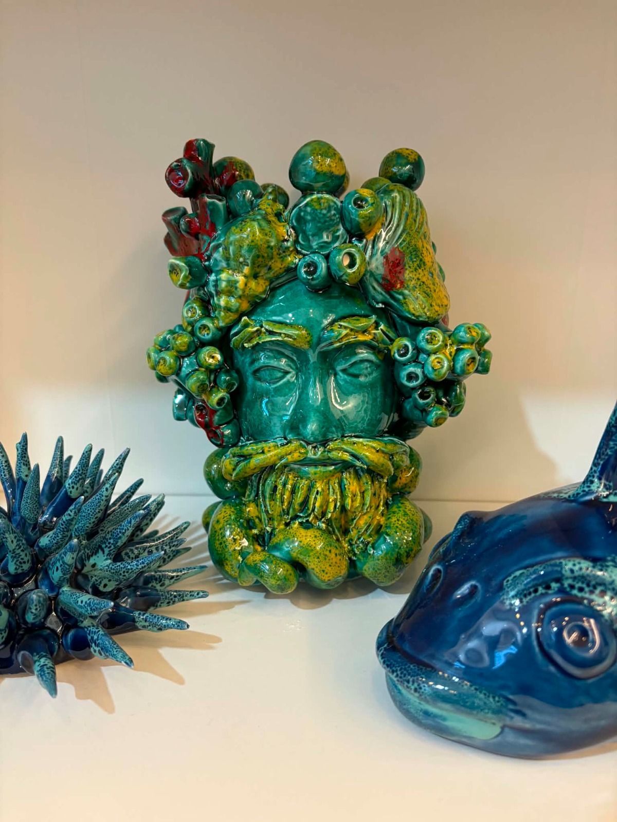 Tre sculture in ceramica color turchese e verde: due vasi a forma di viso e una pigna su una mensola bianca.