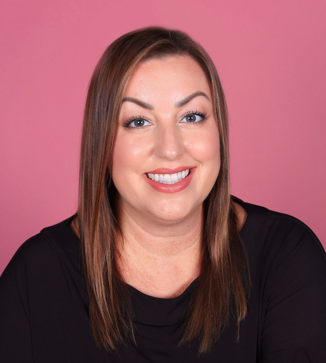 Woman with light brown hair smiles, wearing black shirt against a pink background.