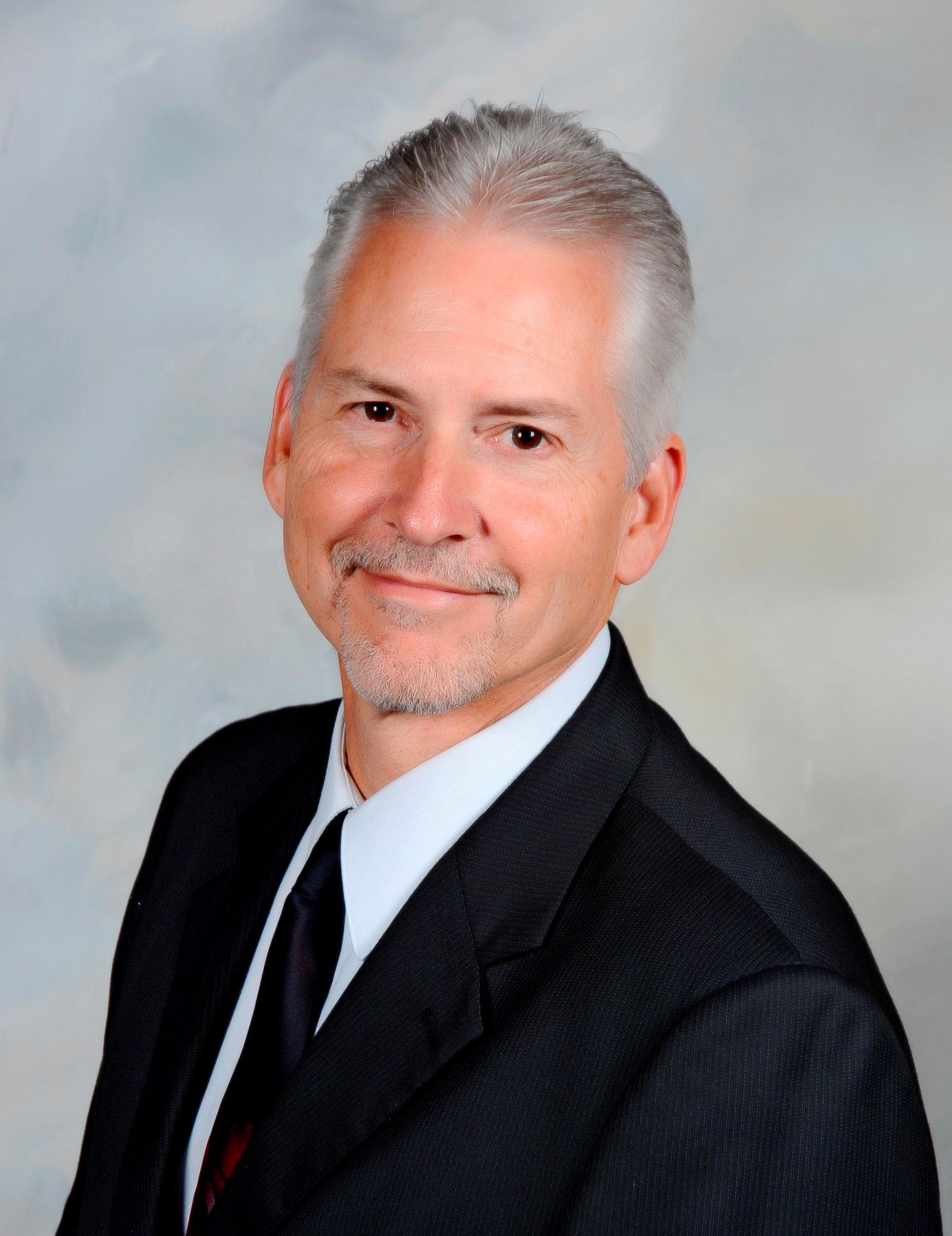 Man in a suit smiles at the camera, against a soft, light blue and gray background.
