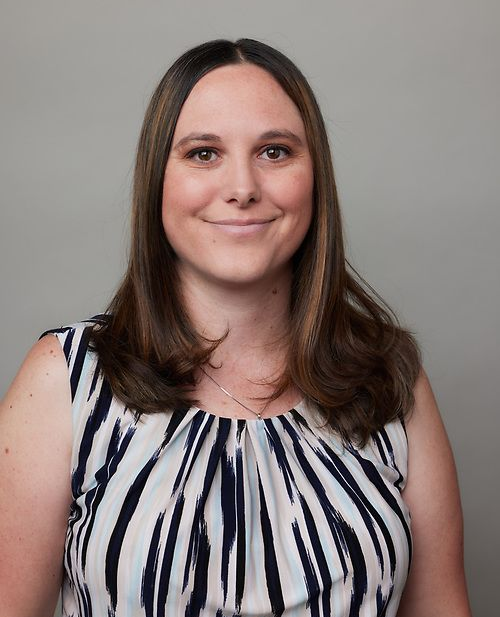 Woman with long brown hair, wearing a navy and white striped sleeveless top, smiles.