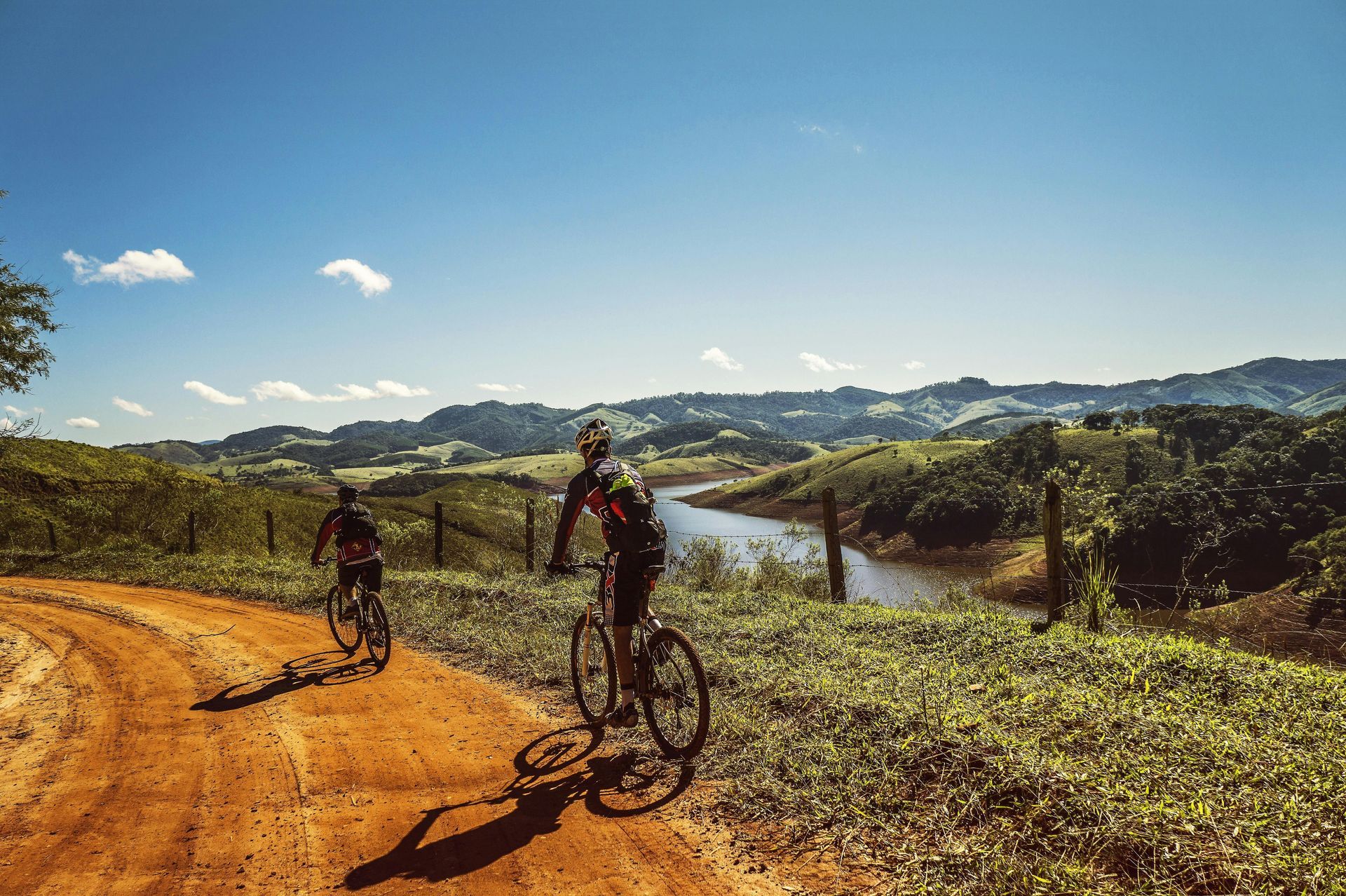 Two cyclists riding bikes on a dirt path with a lake and hills in the background on a sunny day.