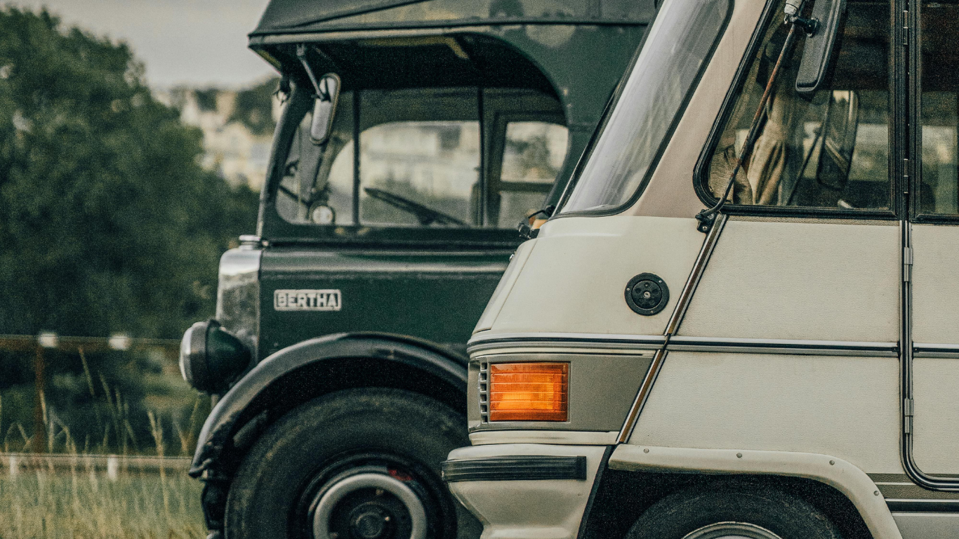 Two vintage vehicles, dark green bus and white van, parked side-by-side in an outdoor setting.