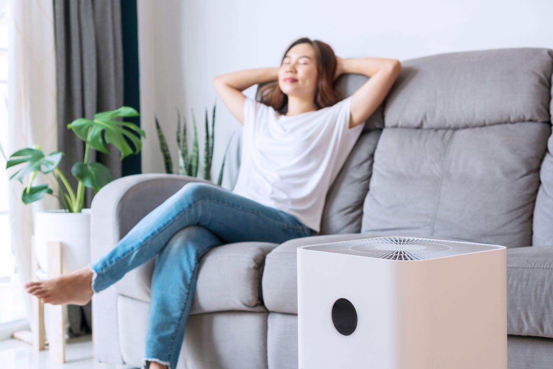 A woman is sitting on a couch next to a air purifier.