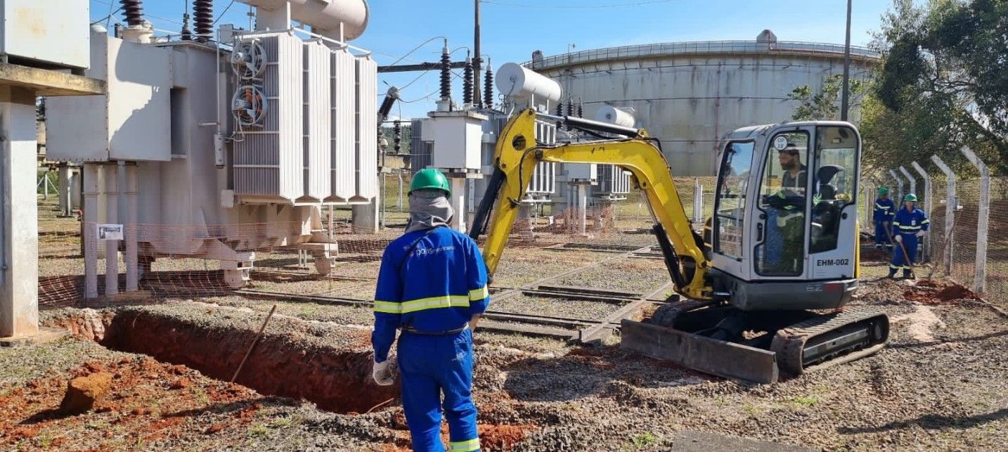 Um homem de macacão azul está parado em frente a uma escavadeira amarela.