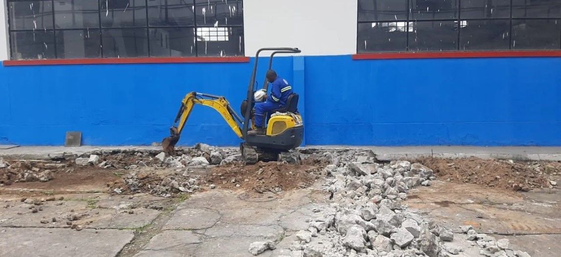 Um homem está dirigindo uma pequena escavadeira amarela em uma estrada de terra em frente a um prédio azul e branco.