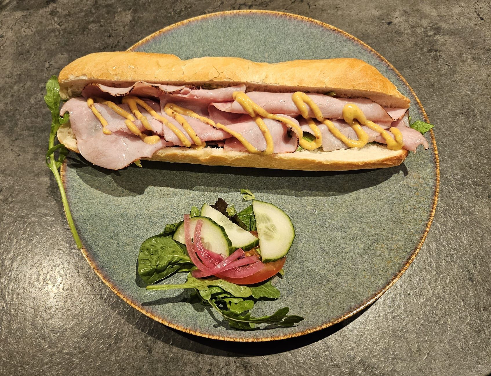 Ham sandwich met mosterd op een bord, met een bijgerecht van salade met komkommer en ingelegde uitjes.