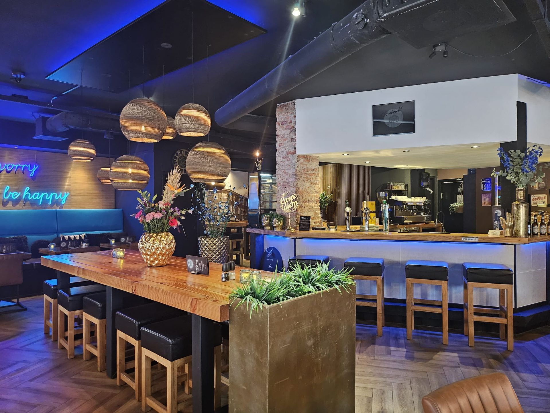 Restaurantinterieur met bar en lange houten tafel, blauwe verlichting en decoratieve hanglampen.