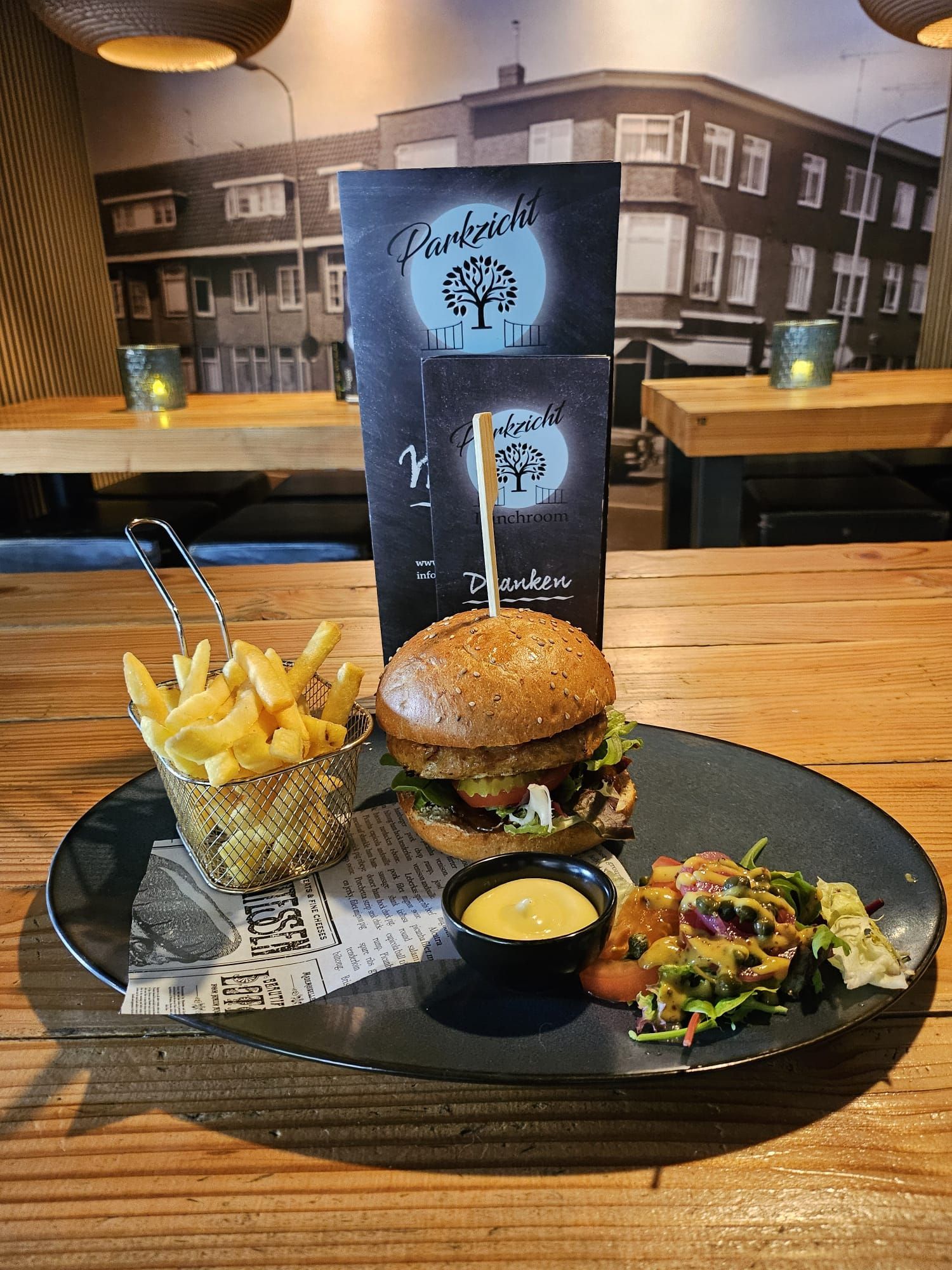 Een hamburger, friet en salade op een bord, met menukaarten op de achtergrond, op een houten tafel.