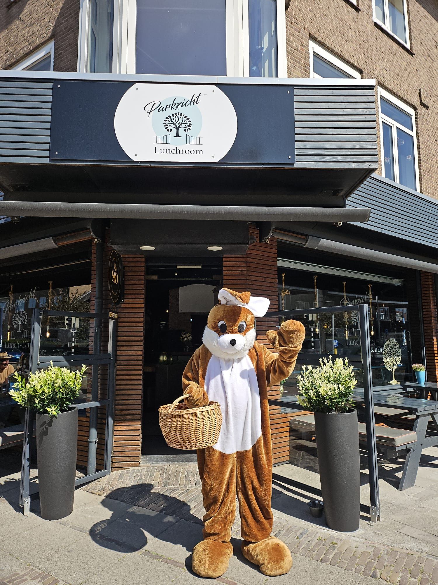 Een persoon in een konijnenpak zwaait met een mandje in zijn hand voor de ingang van een restaurant.