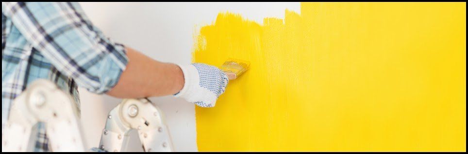 A person paints a wall yellow with a roller, wearing a plaid shirt and gloves.