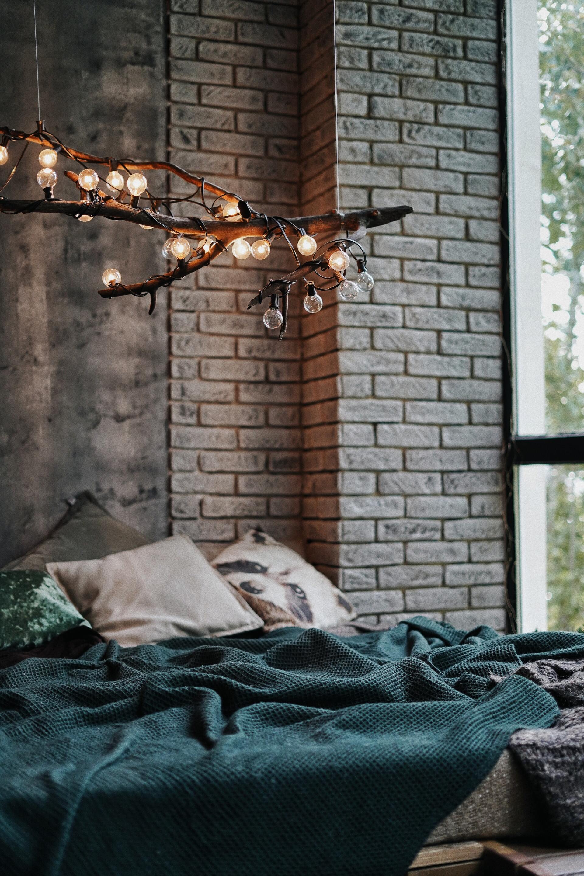 Bed with teal blanket and pillows, against brick wall and window. Branch chandelier with lights.