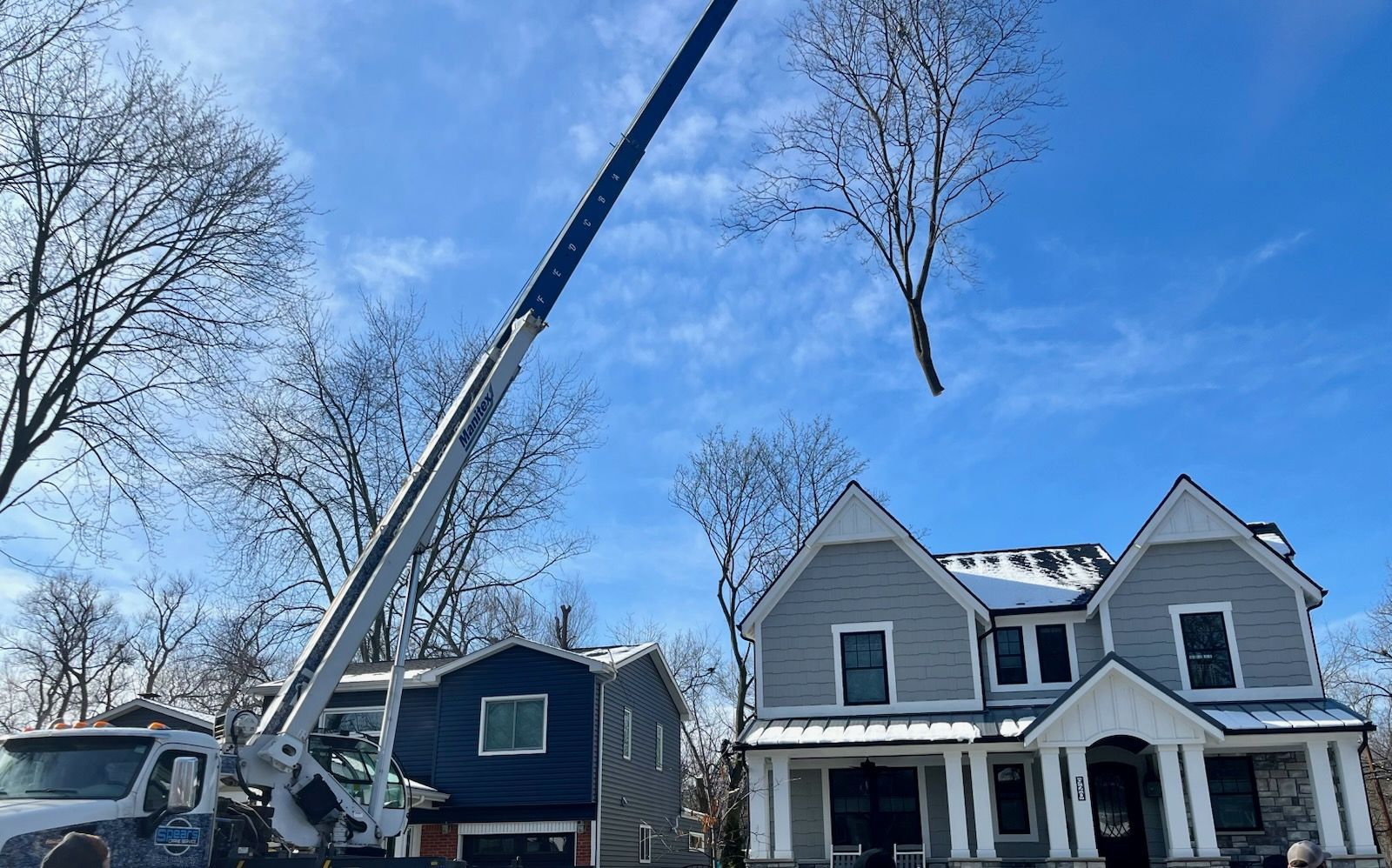 Truck with lift trimming a tree in front of a house. Other houses and a green machine are also visible.