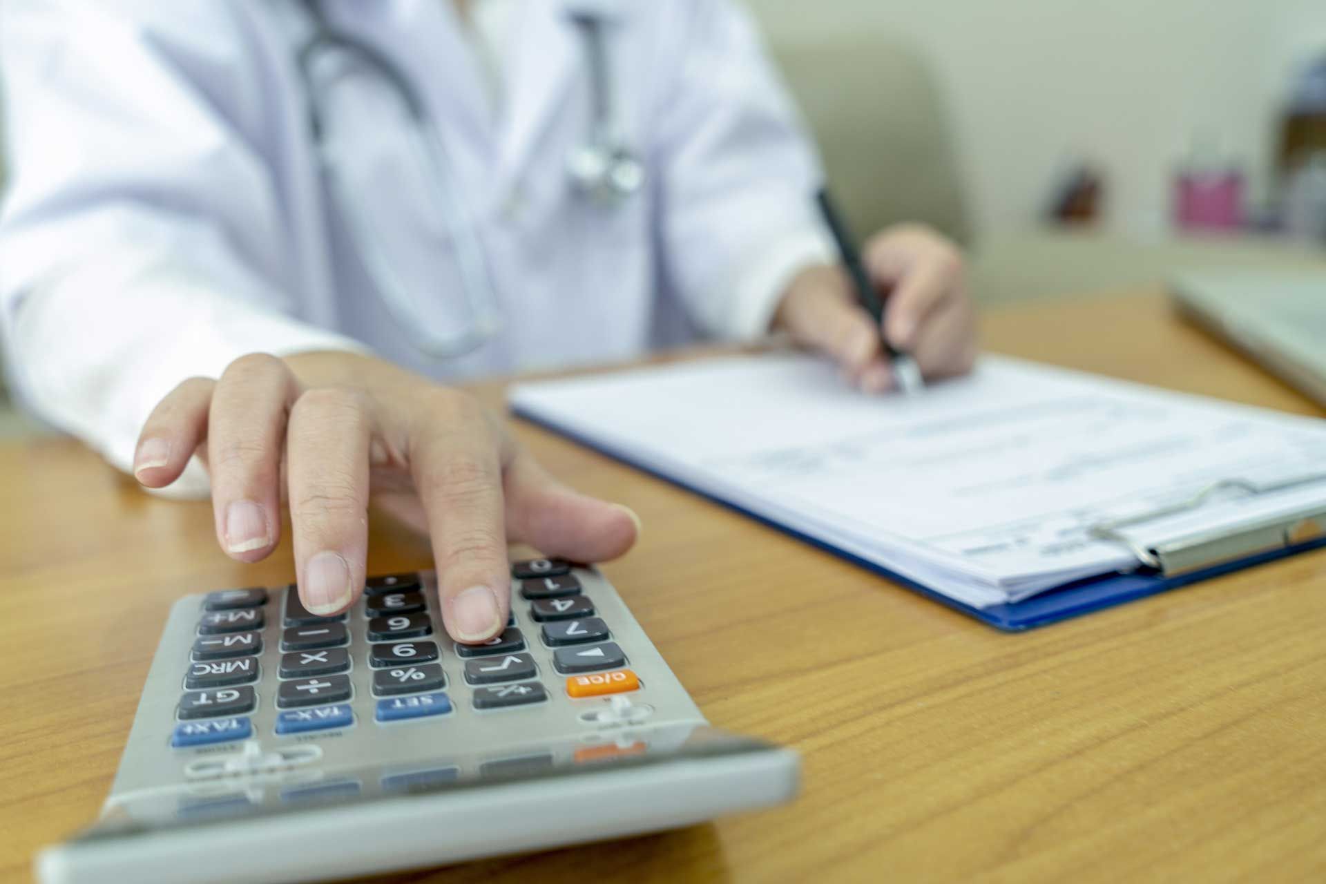 Doctor in white coat uses calculator, writing on clipboard at desk.