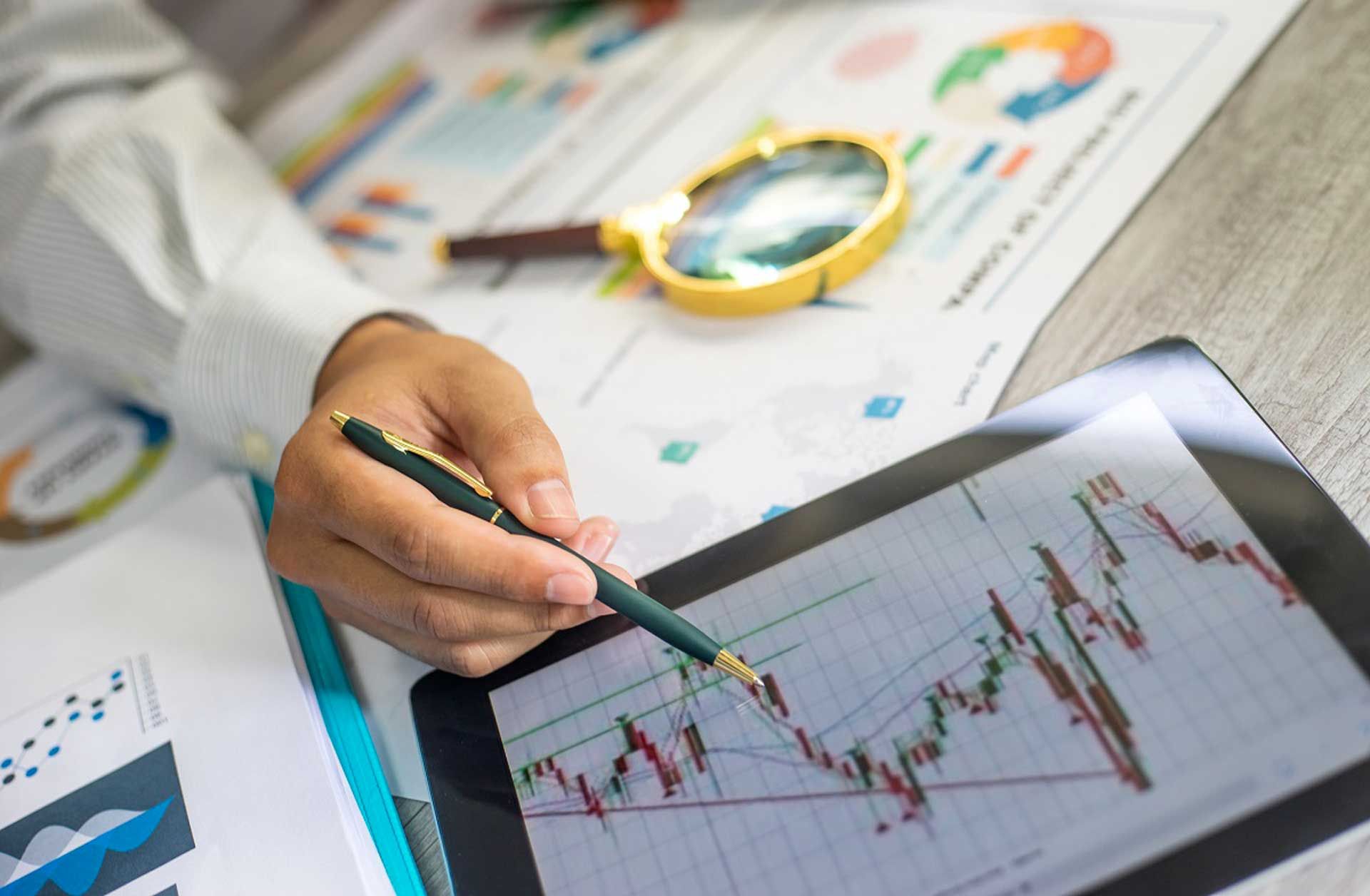 Person analyzing financial charts on a tablet, with papers, pen, and magnifying glass on a desk.