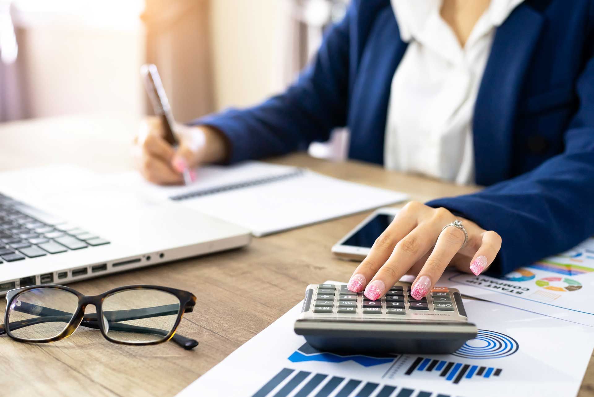 Woman in blue blazer calculating with a calculator at desk with laptop, phone, and financial graphs.