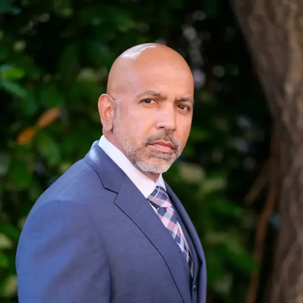 A bald man in a suit and tie is standing in front of a tree.