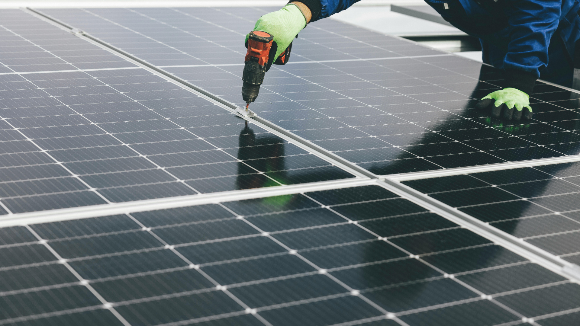 Person installing solar panels on a roof, using a power drill, wearing gloves.