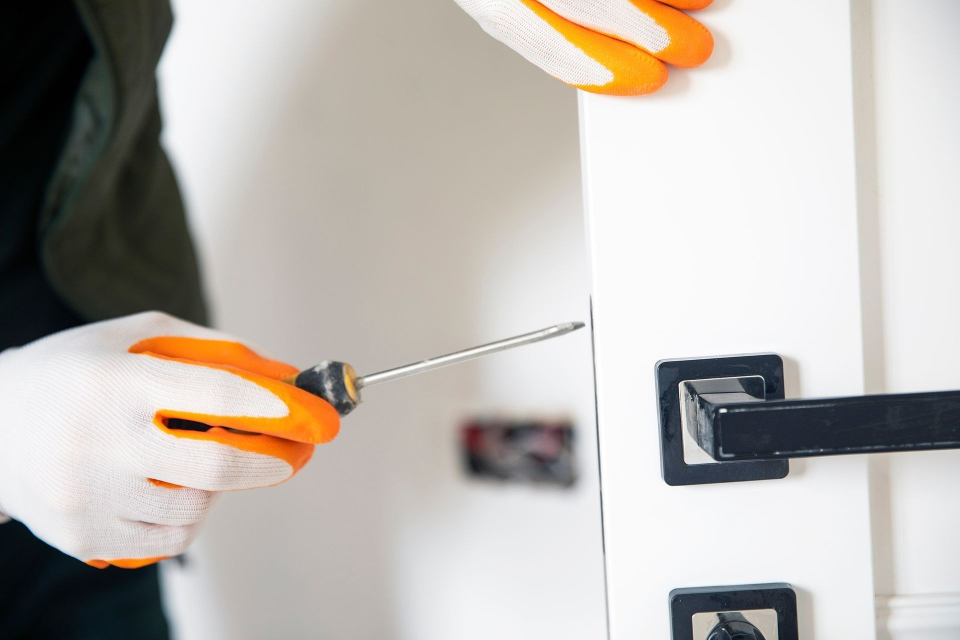 Gloved hands use a screwdriver on a white door near a black handle.