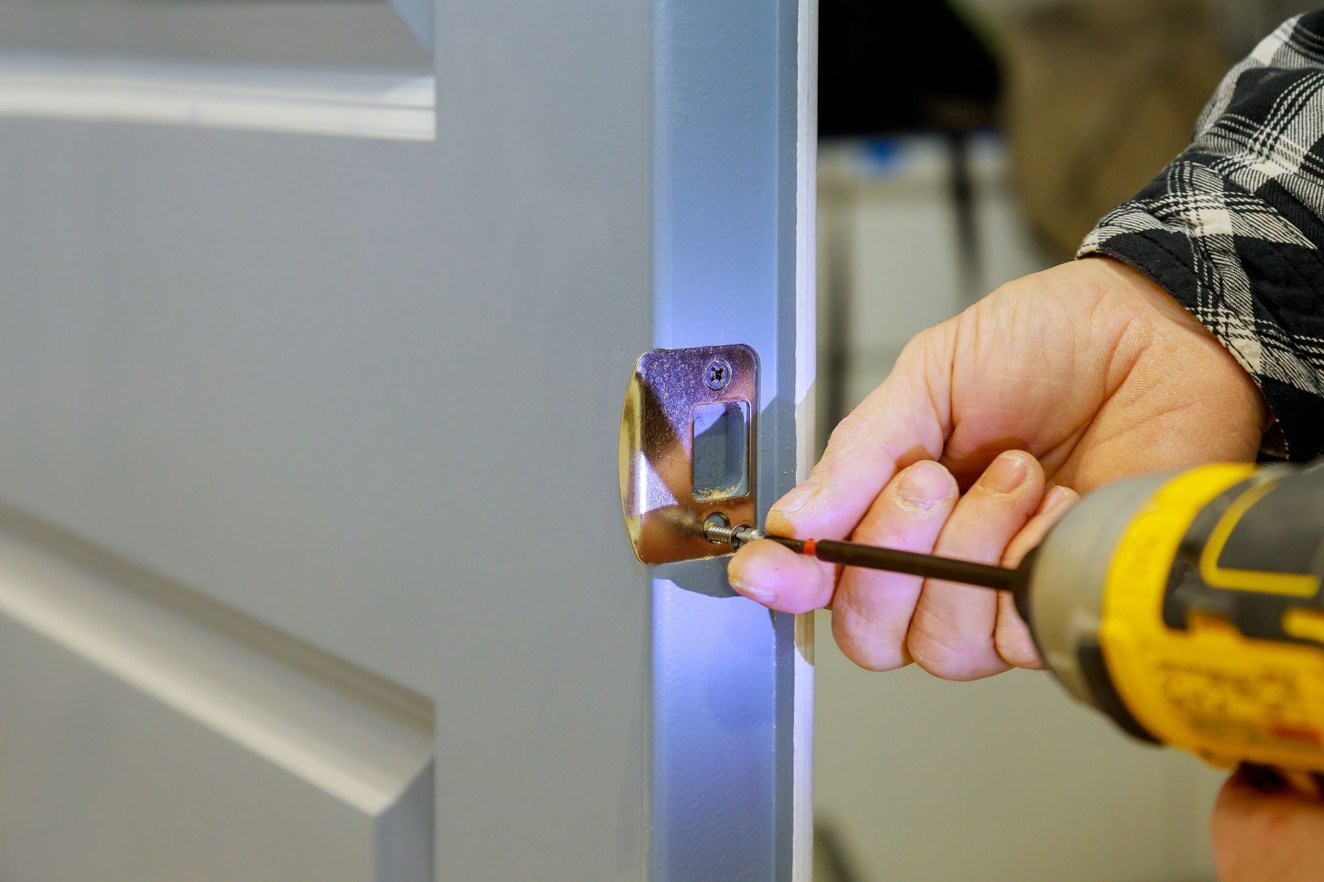 A close-up of a person using a power drill to screw a metal door latch plate into the edge of a grey door.