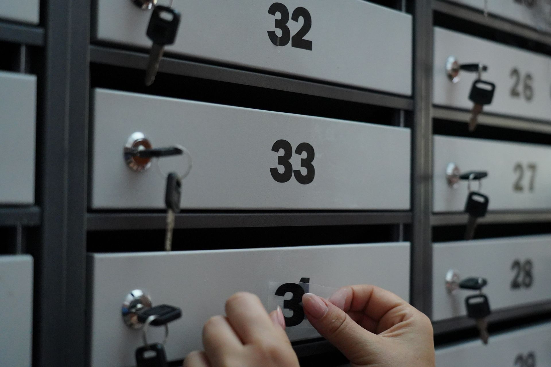 A person’s hands peel a black number label off a grey mailbox door numbered 33 in a row of mailboxes.