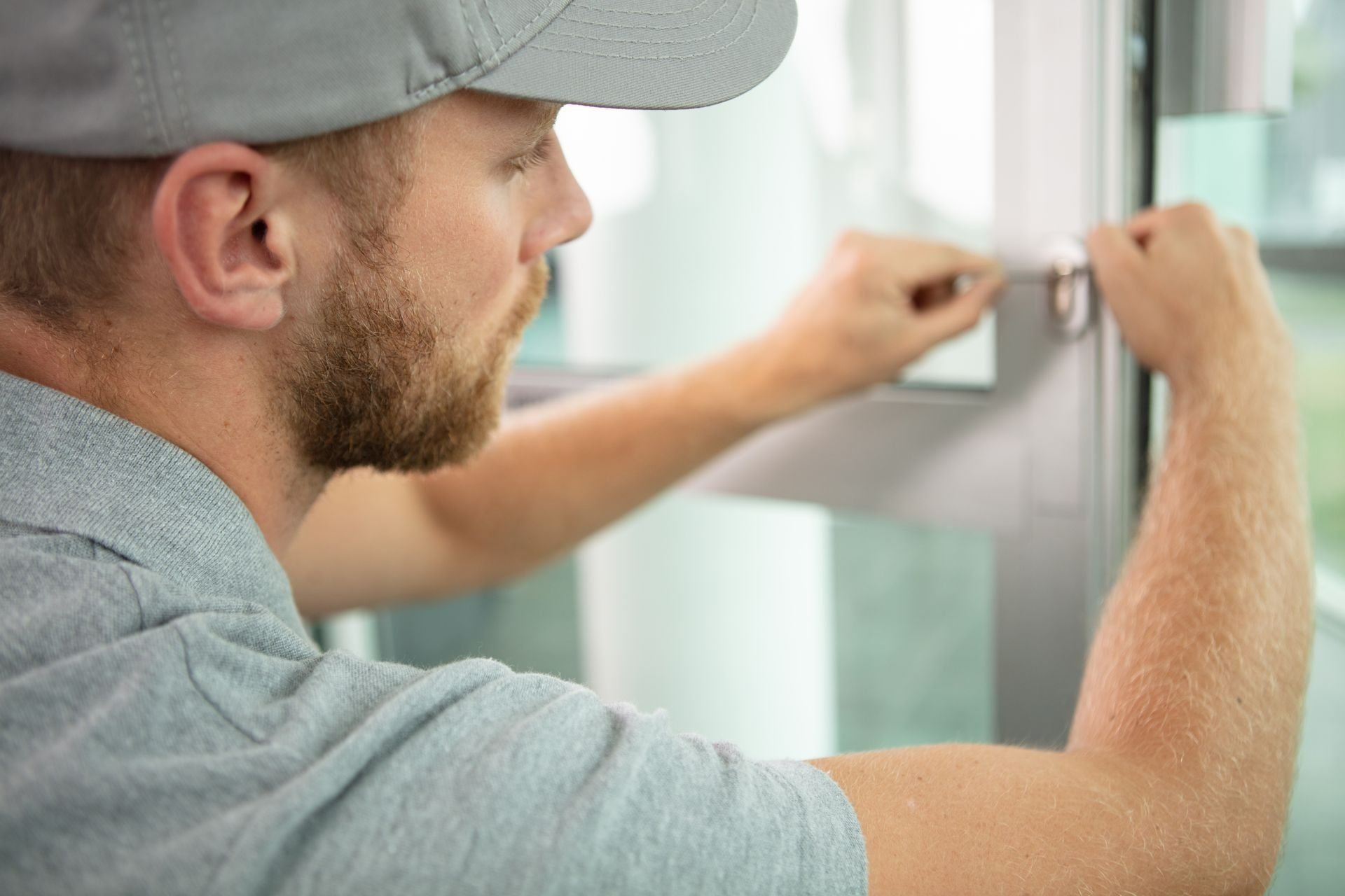 A person wearing a grey cap uses a small tool to adjust a metal window frame.