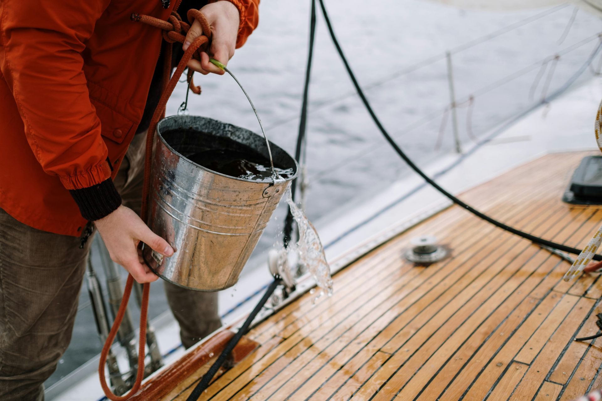 Person in red jacket emptying water from a metal bucket onto a wooden boat deck.