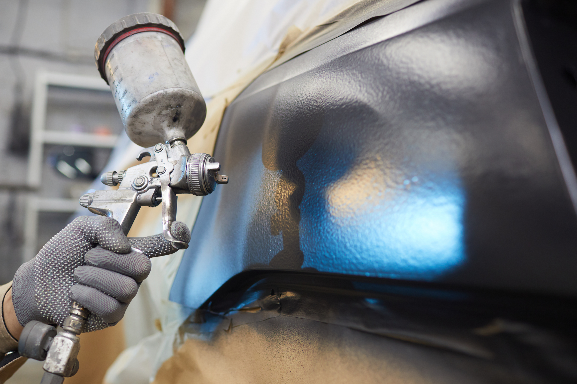 Person spray painting a black car bumper with a silver spray gun, in a repair shop.