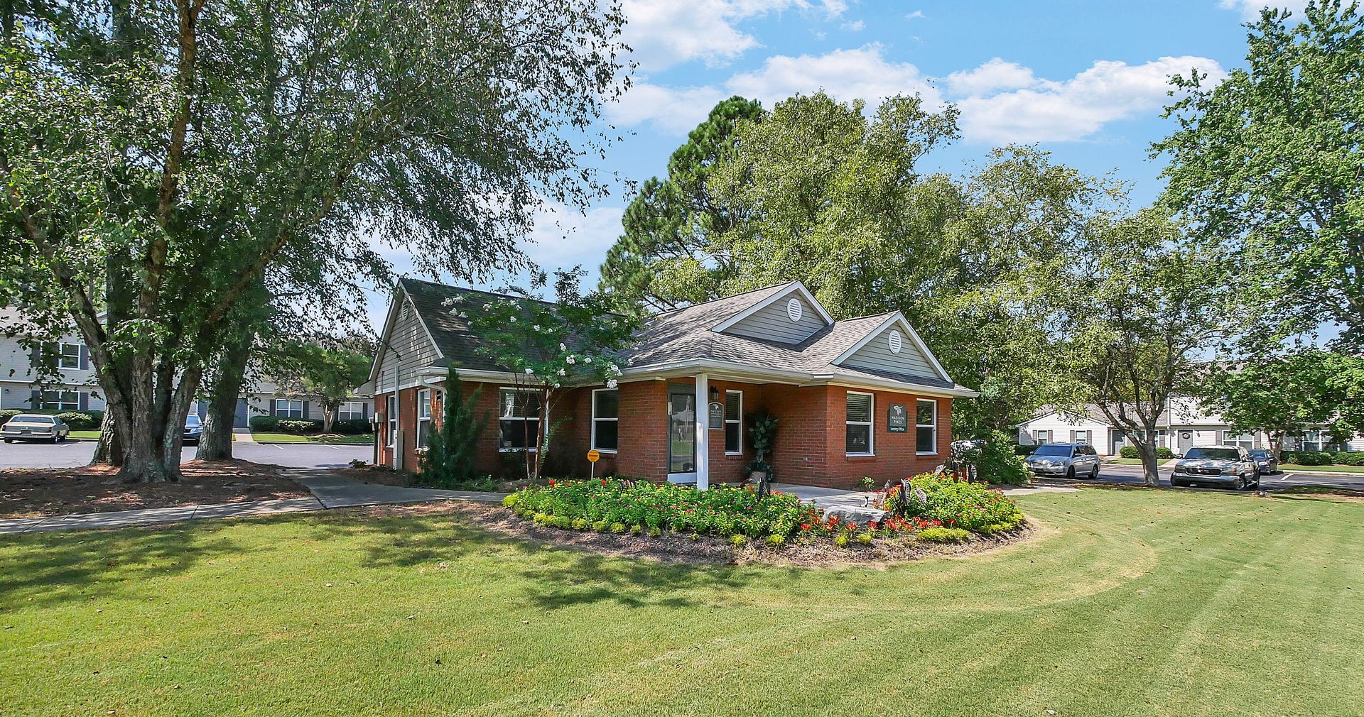 A single-story brick building with a gray shingle roof, surrounded by lush green trees and a well-maintained lawn.