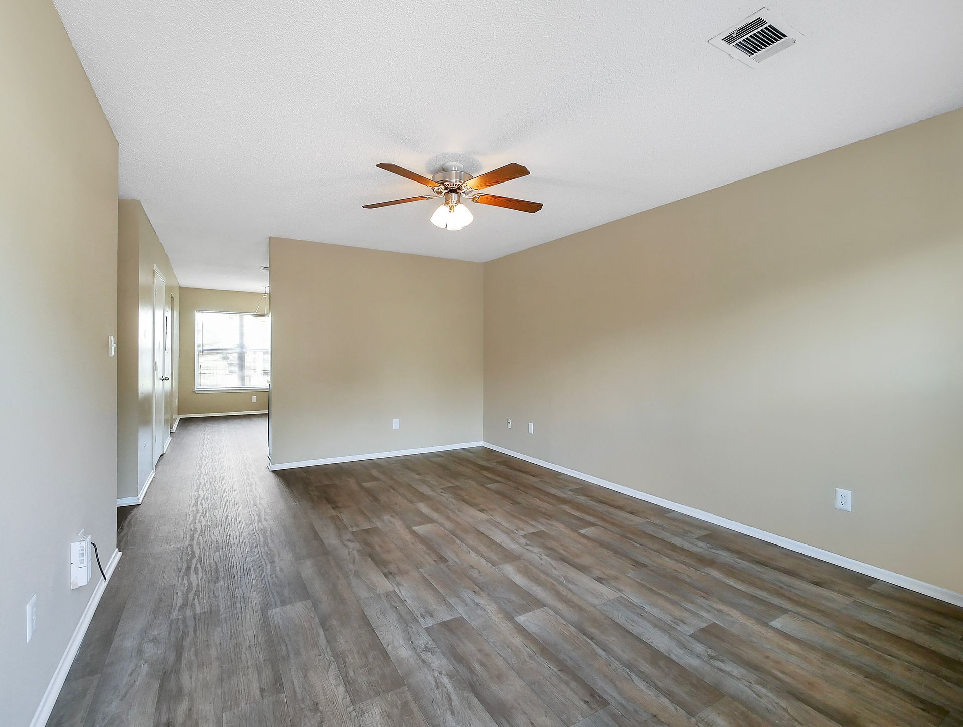 A brightly lit, empty living room with light beige walls, wood-look flooring, and a ceiling fan.