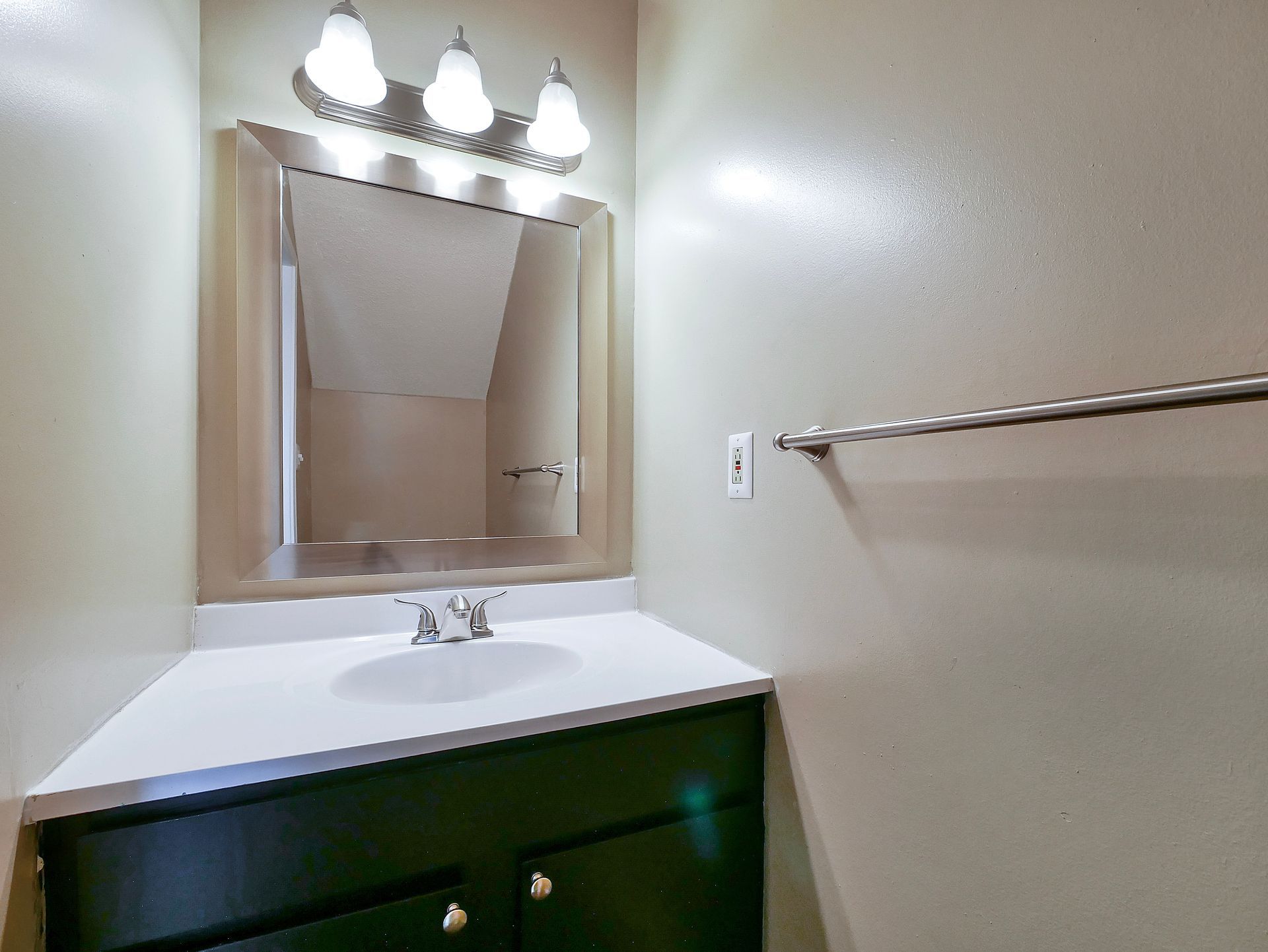 A bathroom vanity with a dark green cabinet, white countertop, sink, square mirror, and a three-light fixture above it.