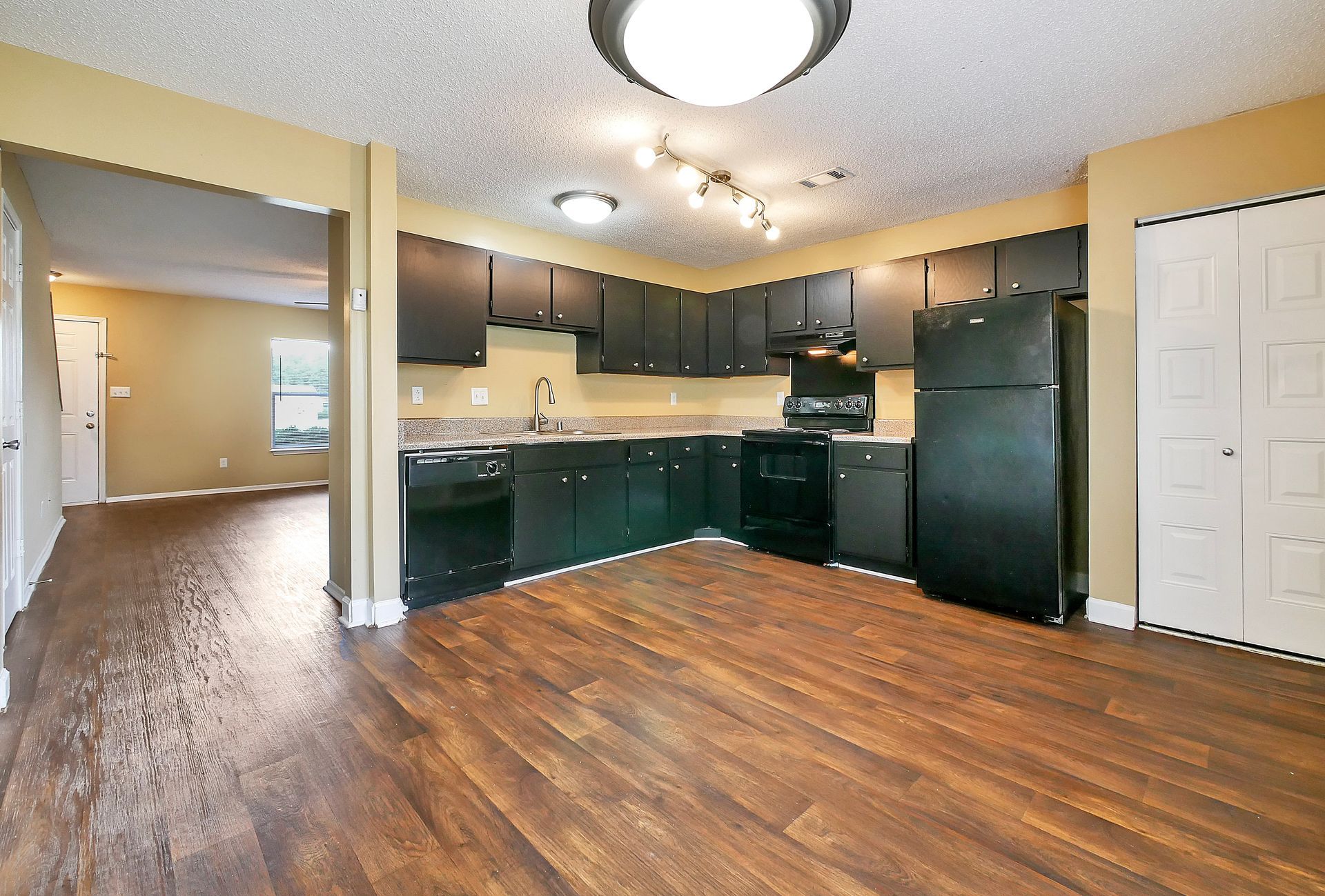 A kitchen with dark cabinets, black appliances, wood-look flooring, and recessed lighting, leading to a living area.