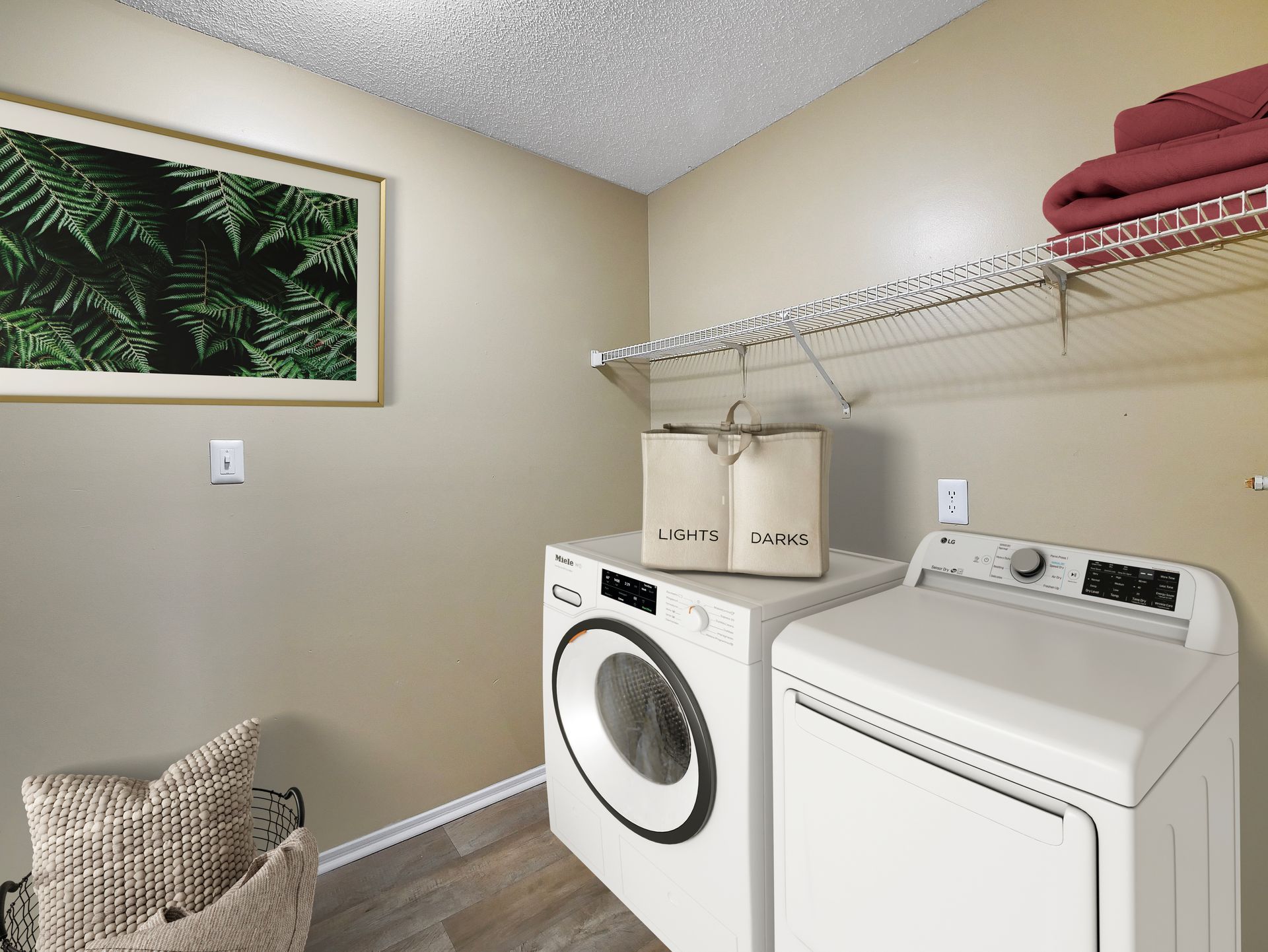 A laundry room with a white washer and dryer, a shelf holding folded towels, and a framed fern print on the tan wall.