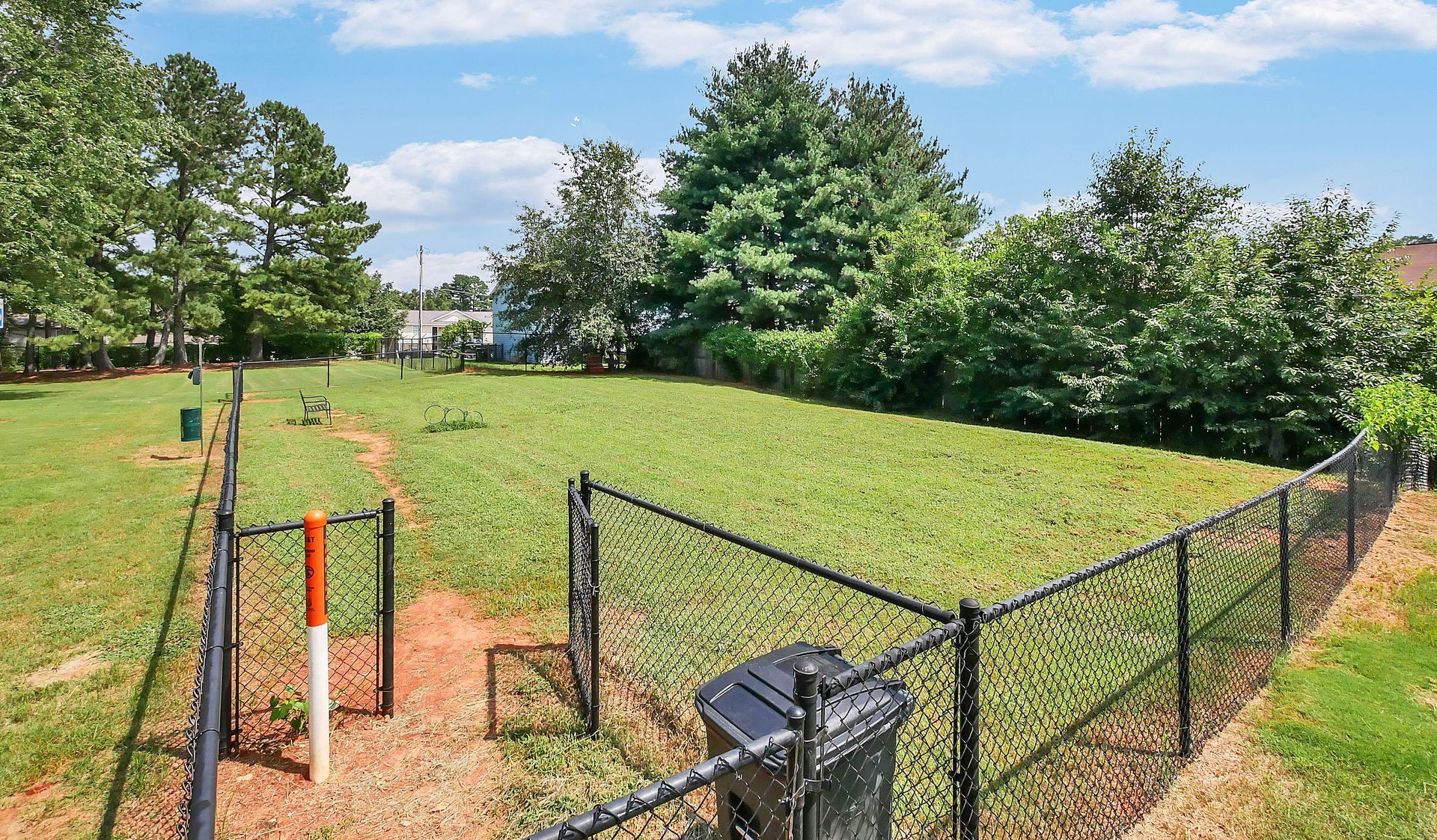 An enclosed, grassy outdoor dog park with a chain-link fence, a gated entrance, and a small black trash bin.