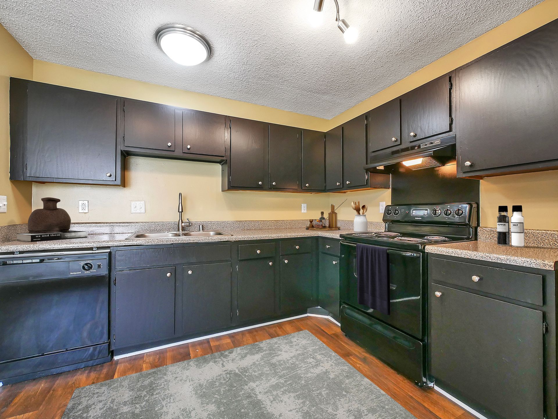 A kitchen with dark cabinets, a speckled countertop, a black dishwasher, and a stove under an overhead light.