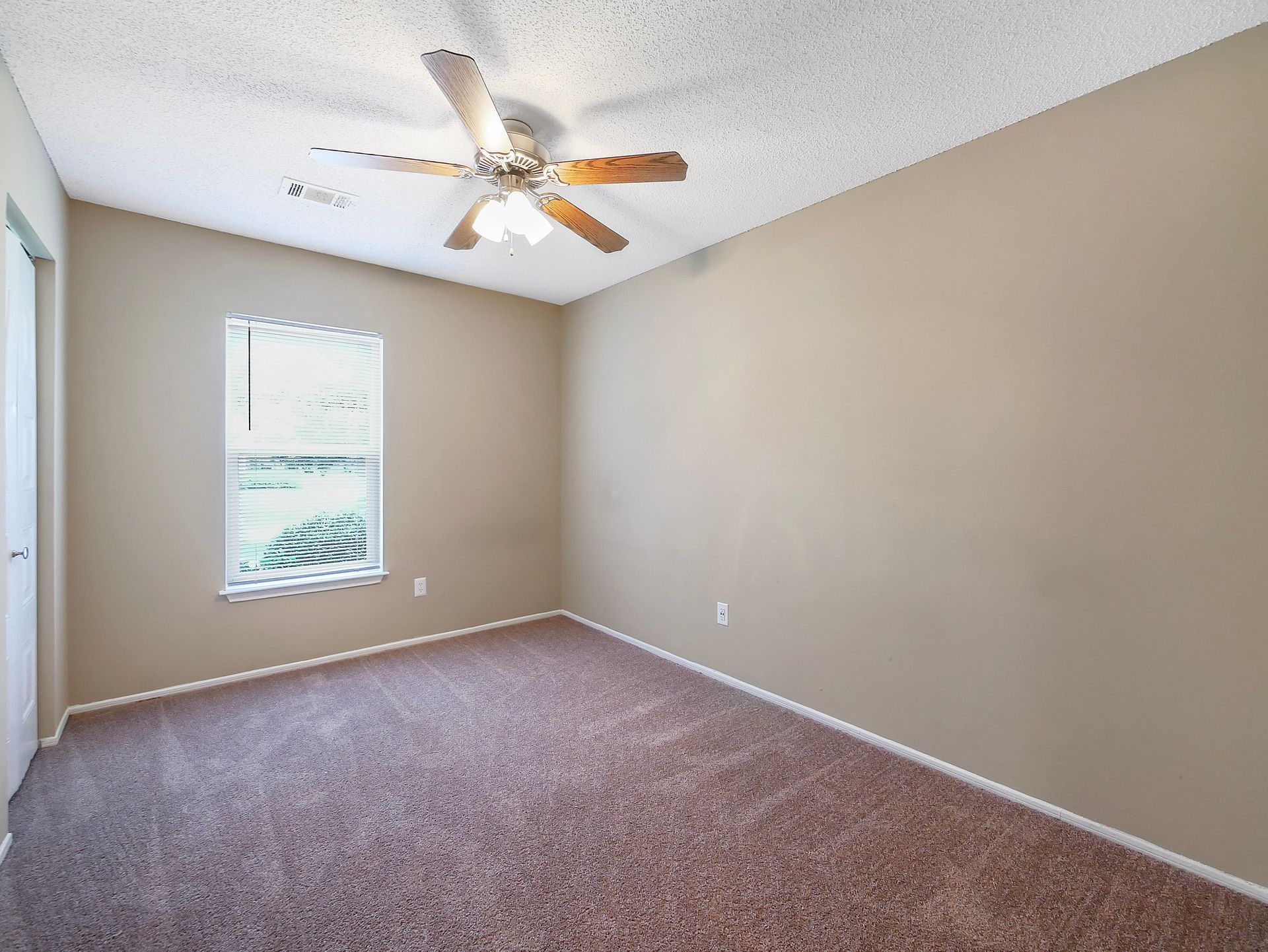 An empty bedroom with tan walls, brown carpet, a ceiling fan, and a window with blinds on the left wall.
