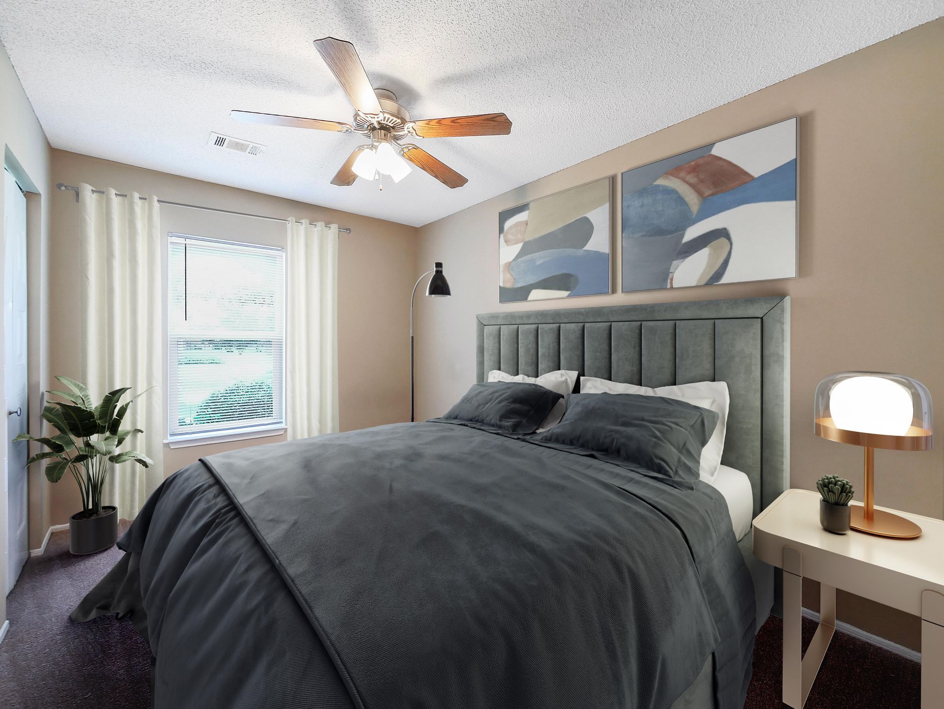 A modern bedroom featuring a grey tufted headboard, a dark grey bed, a ceiling fan, and two abstract art pieces.