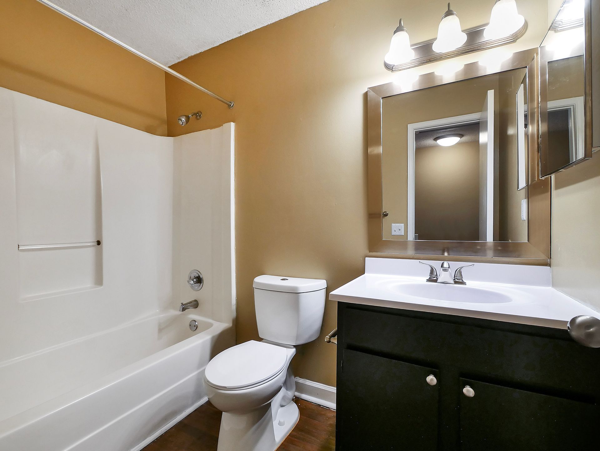A bathroom with tan walls, a white bathtub and toilet, and a vanity cabinet with a mirror and light fixture above.
