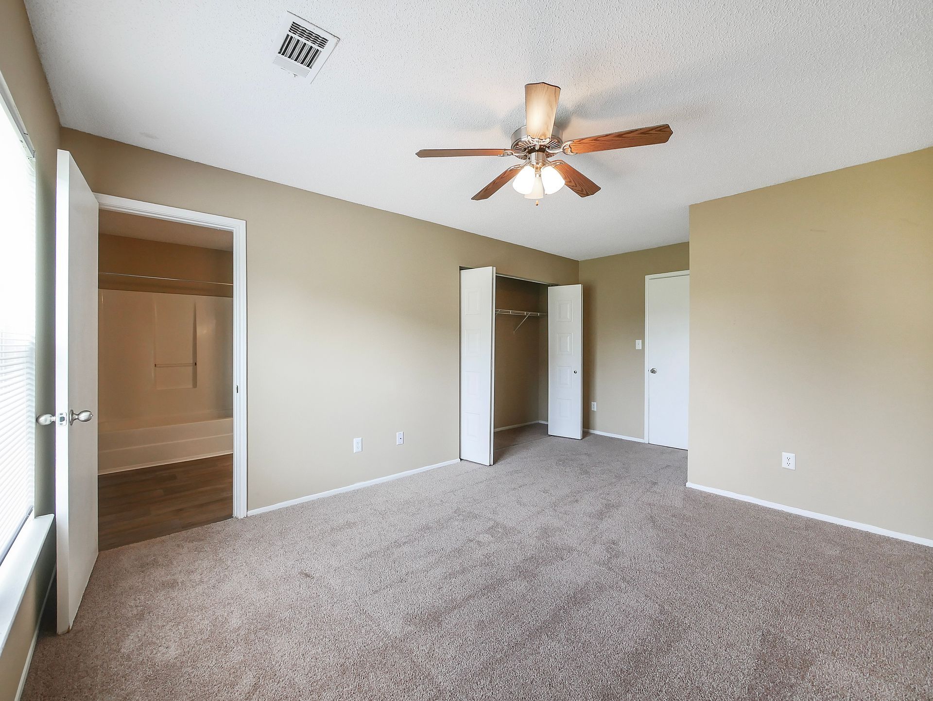 An empty bedroom with tan walls, carpeted flooring, a ceiling fan, a walk-in closet, and an open bathroom doorway.