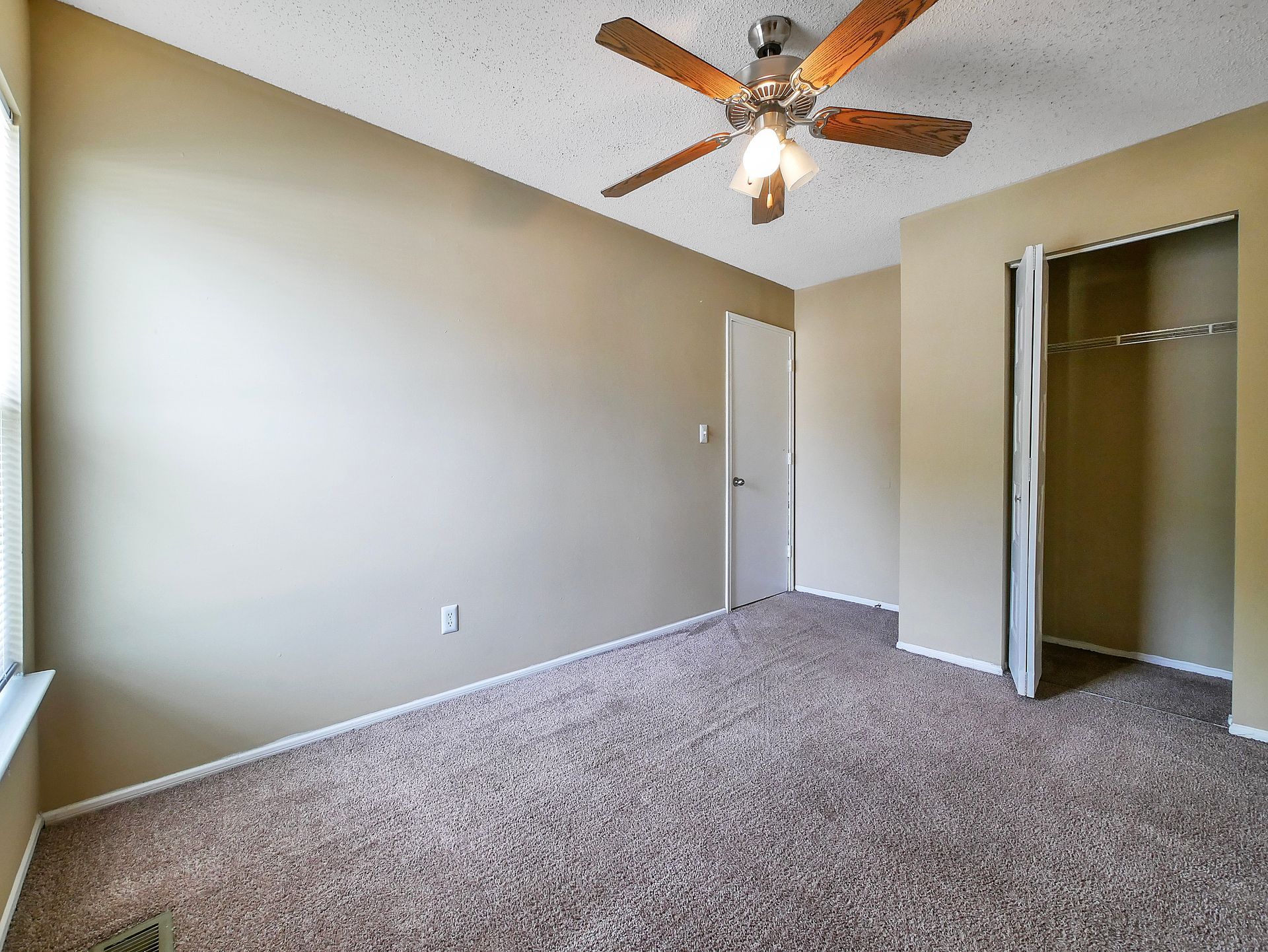 An empty bedroom with tan walls, brown carpet, a ceiling fan, a white door, and an open closet.
