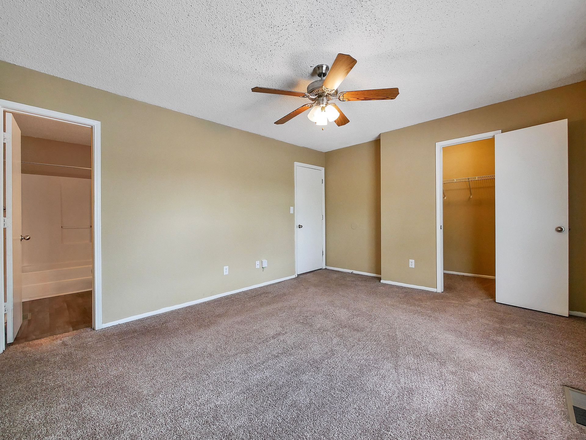 An empty bedroom with tan walls, brown patterned carpet, a ceiling fan, a doorway to a bathroom, and a closet door open.
