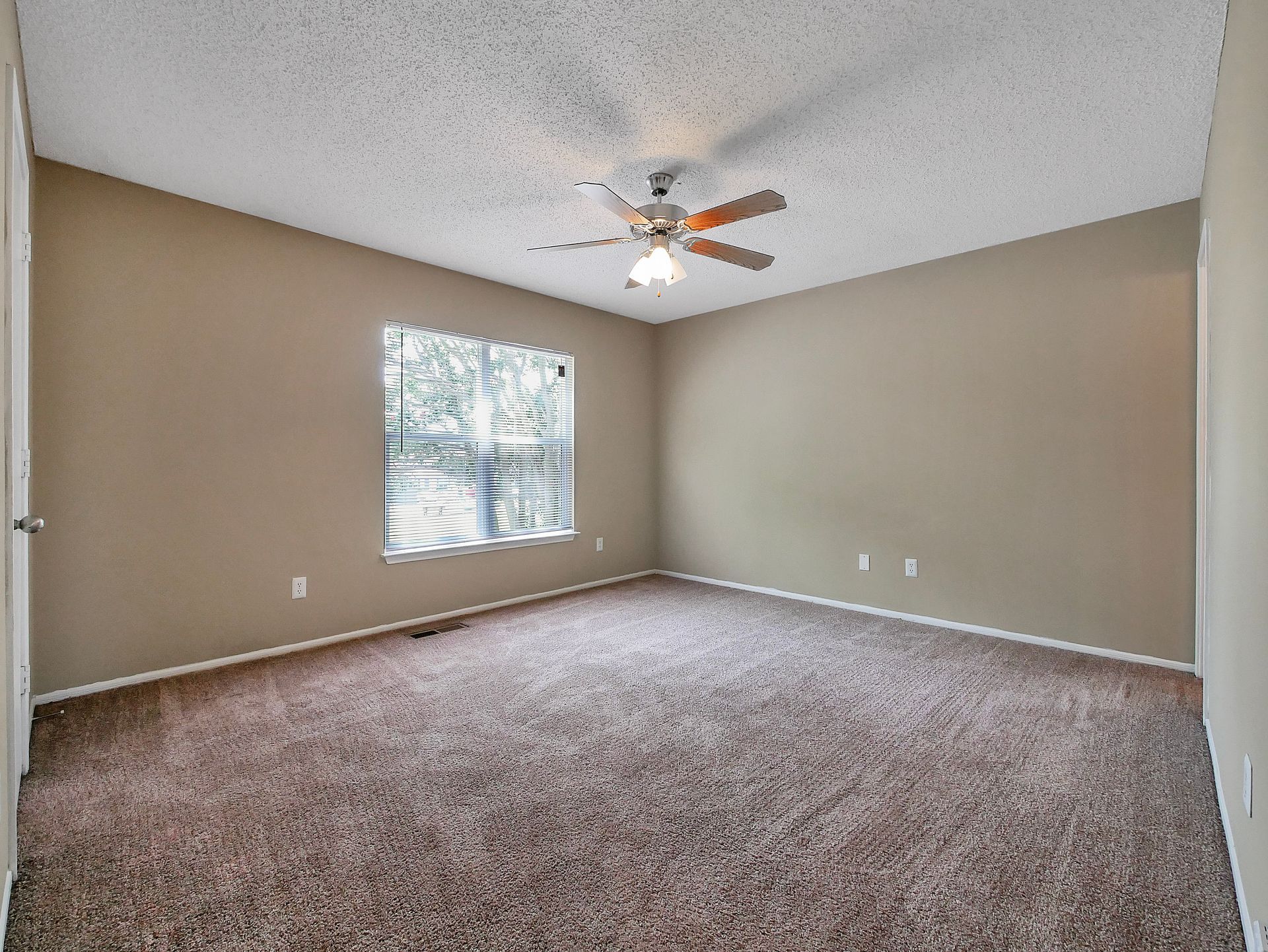 An empty bedroom with tan walls, brown carpet, a ceiling fan, and a window with blinds.