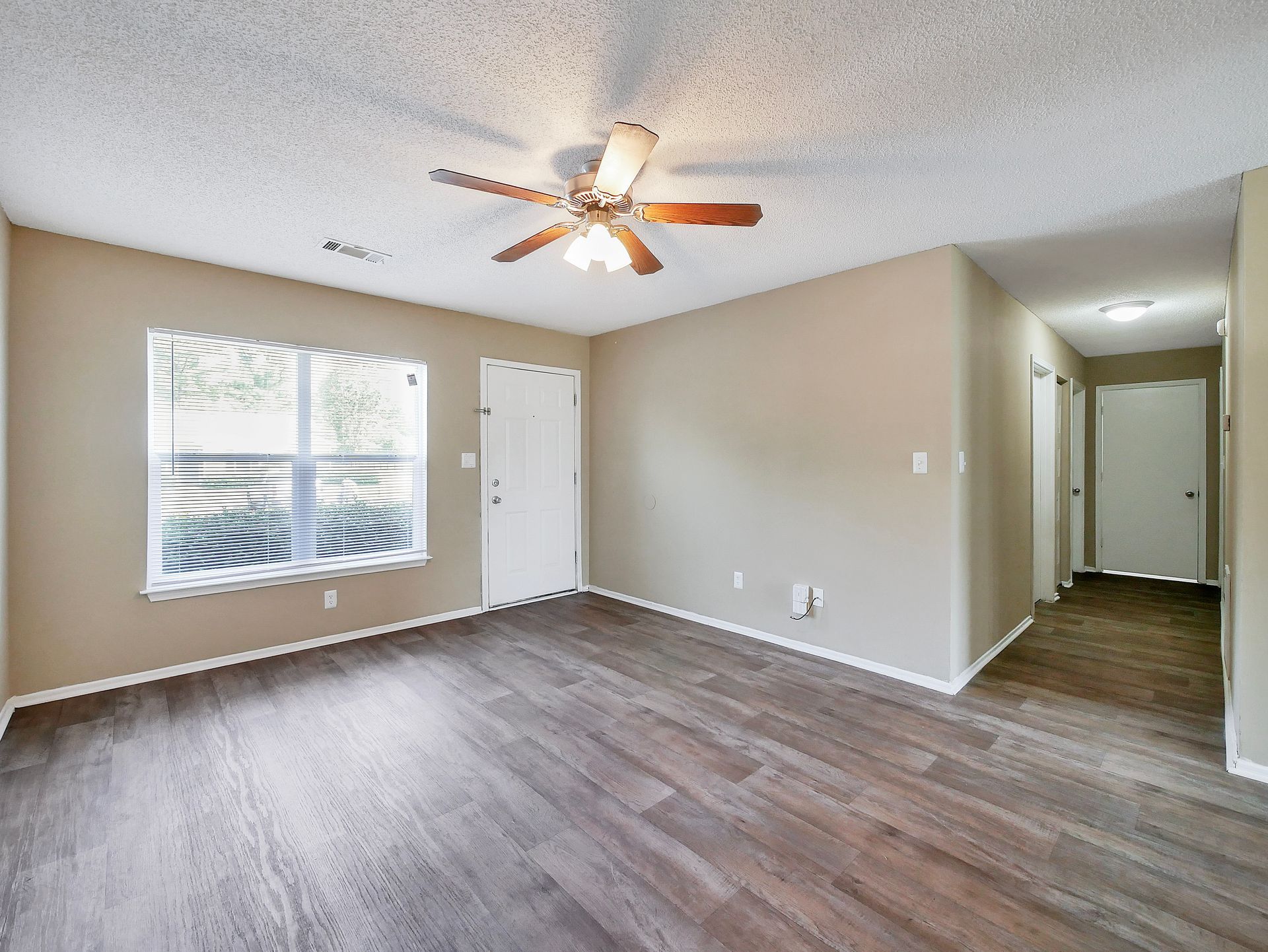 An empty living room with light brown walls, wood-look flooring, a ceiling fan, a window with blinds, and a hallway.